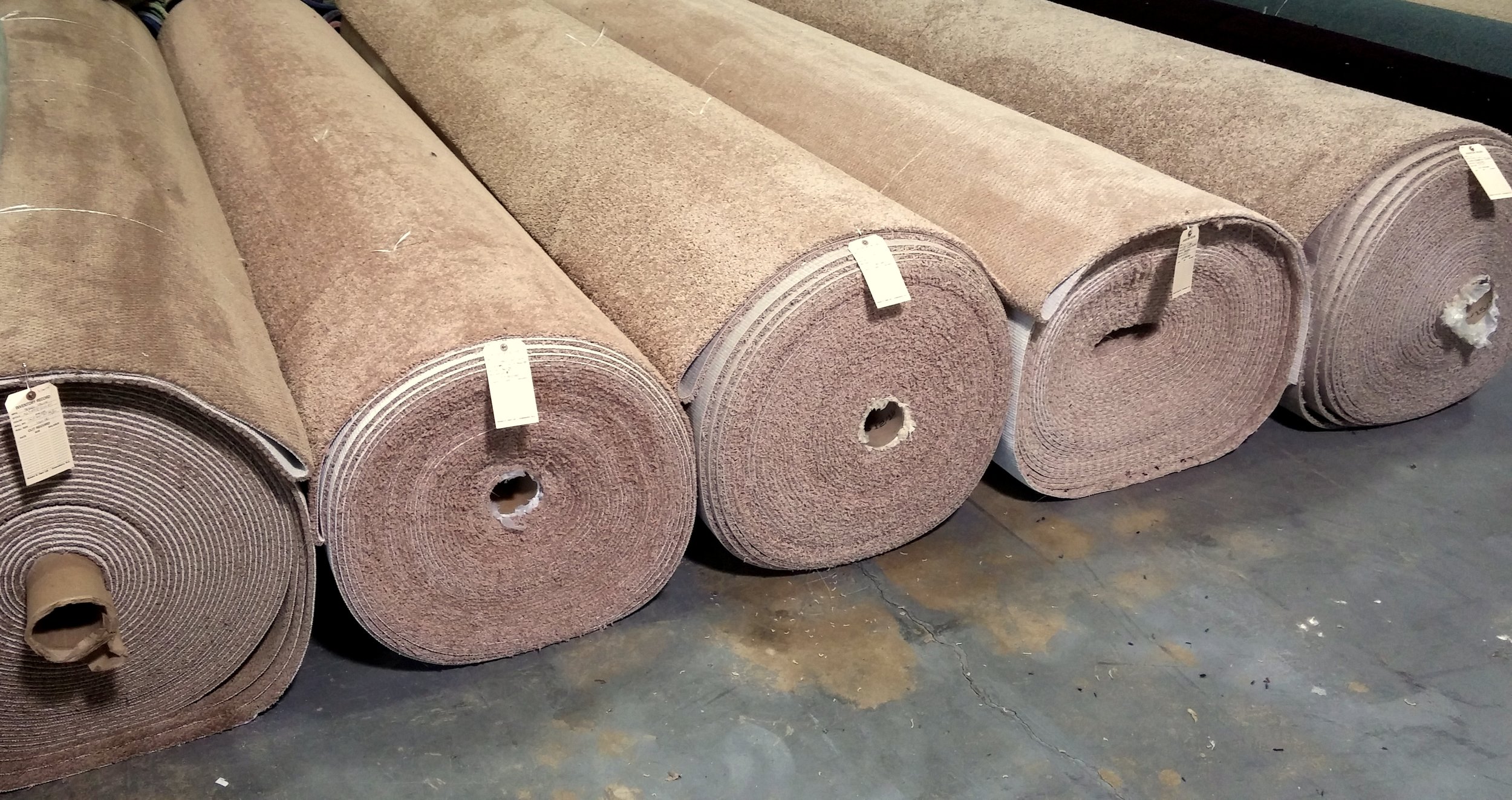 Several large beige carpet rolls with inventory tags lined up on a concrete warehouse floor