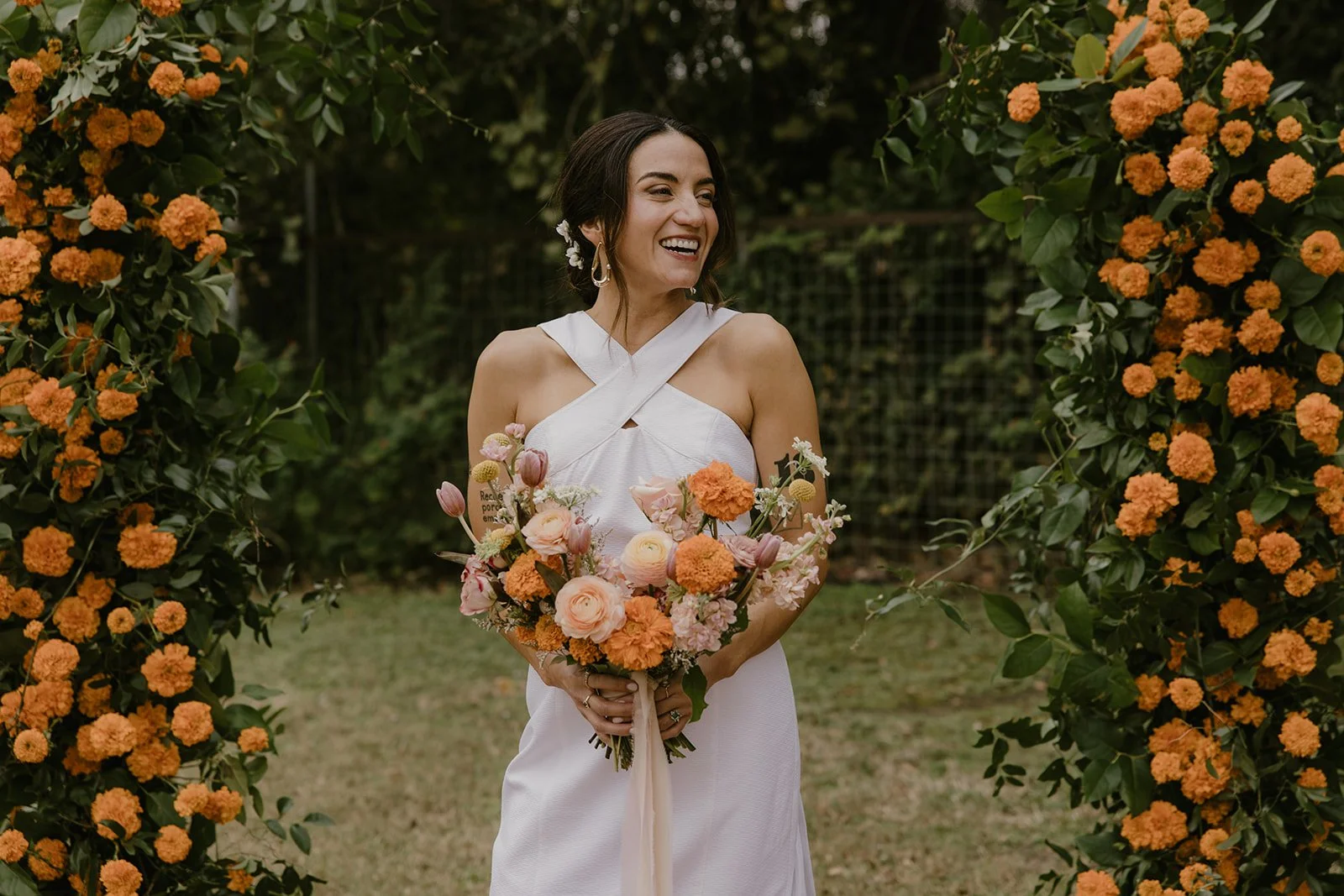 Mexican american bride at Springdale Station surrounded by orange flowers inclusing marigolds. She has a crossed neckline on her dress and large loose tendrils as she looks toward the camera.