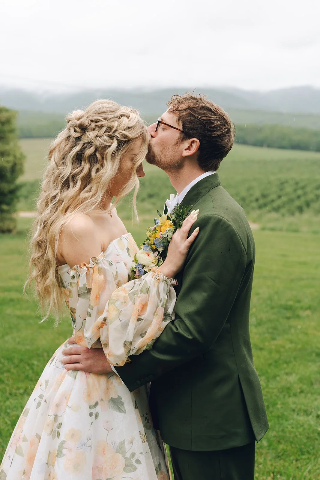 Whimsical couple married on a flower farm with misty mountains in the background. She has a half up hairstyle with a long blonde hair in toussled curls and a dutch 3-strand crown braind.