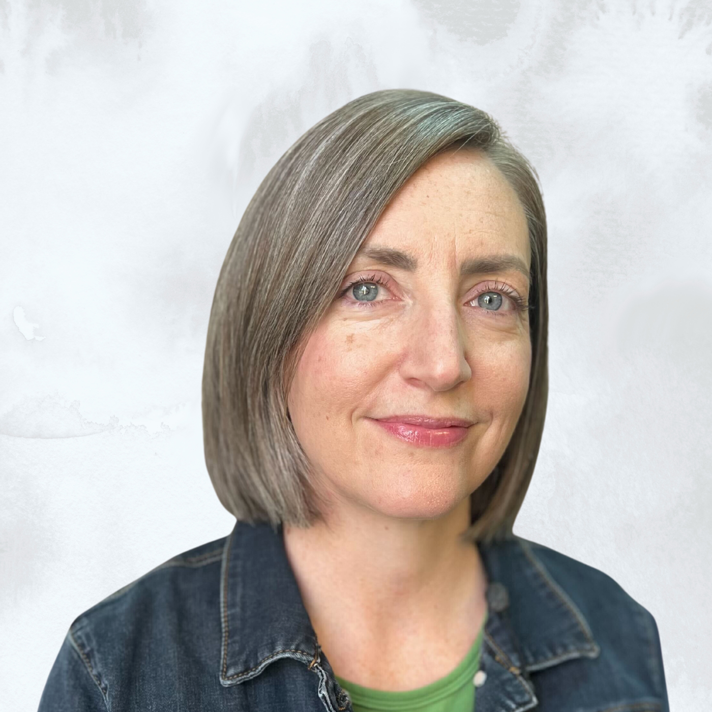 A woman with shoulder-length straight gray hair, blue eyes, and light skin, wearing a dark denim jacket over a green top, smiling softly against a white textured background.