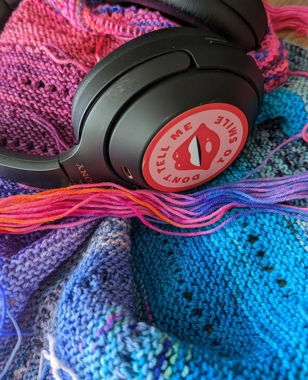 Mmmm colors⁠
.⁠
Description: Over-ear headphones on top of a piece of knitting in pink and blue. The headphones have a sticker on one ear that says &quot;Don't tell me to smile.&quot;