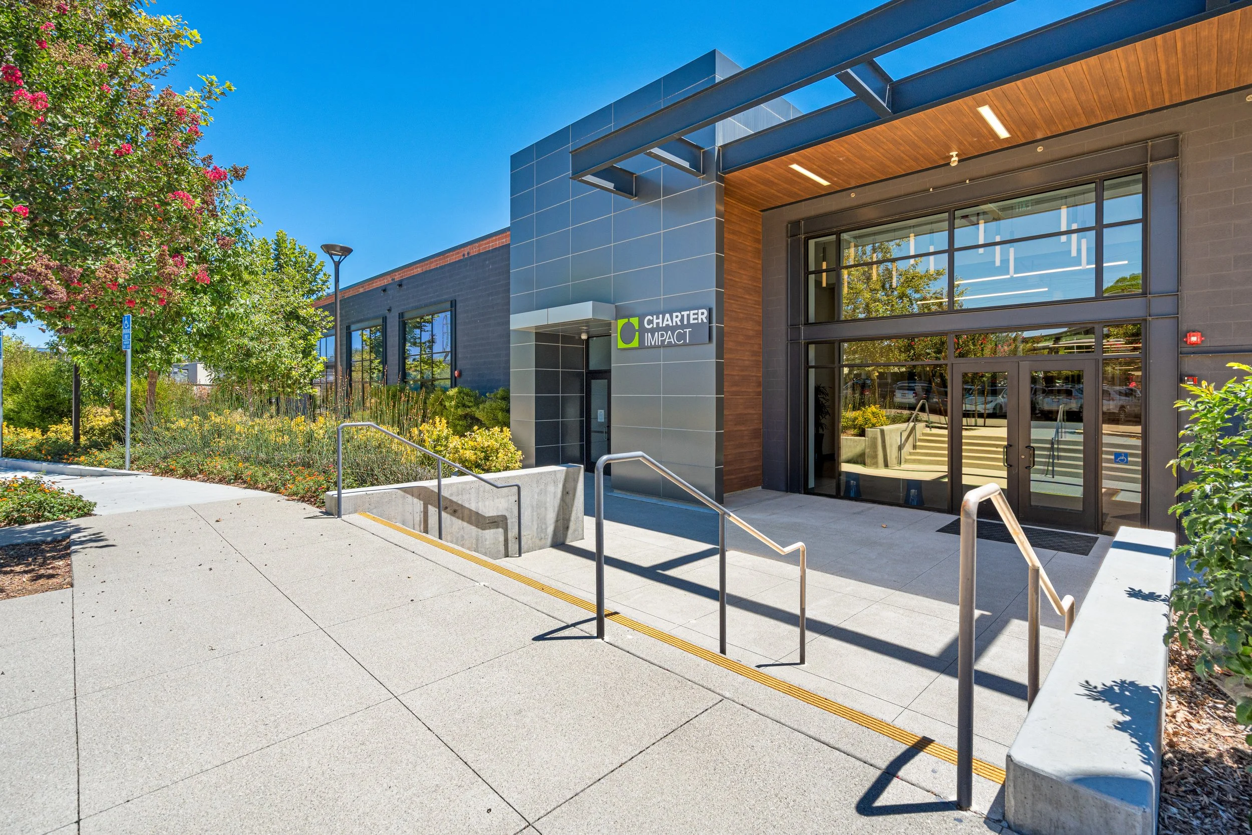 Modern office building with a sign that reads 'Charter Impact' and large glass windows, surrounded by landscaped greenery and a clear blue sky.