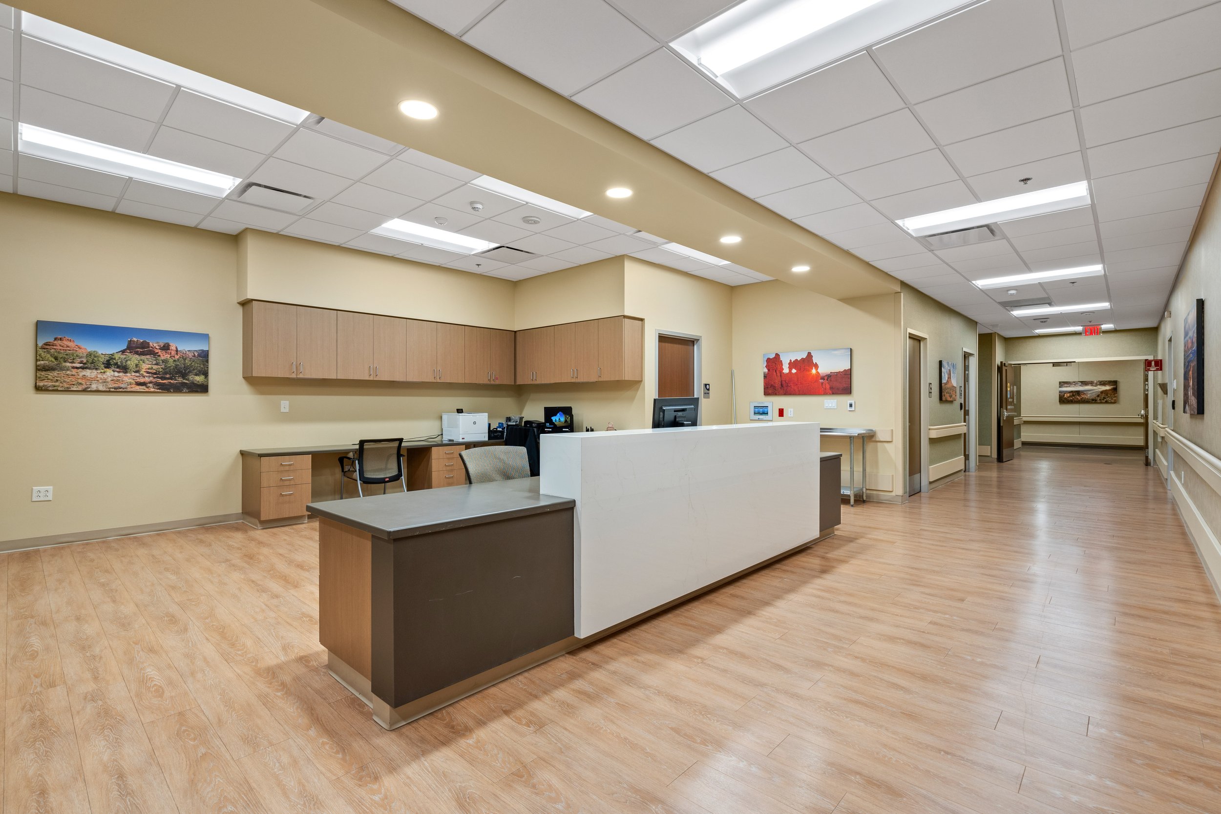 Hospital or clinic reception area with wood floors, beige walls, a white and brown reception desk, a computer, framed landscape photographs on the walls, overhead fluorescent lighting, and a hallway extending into the distance.