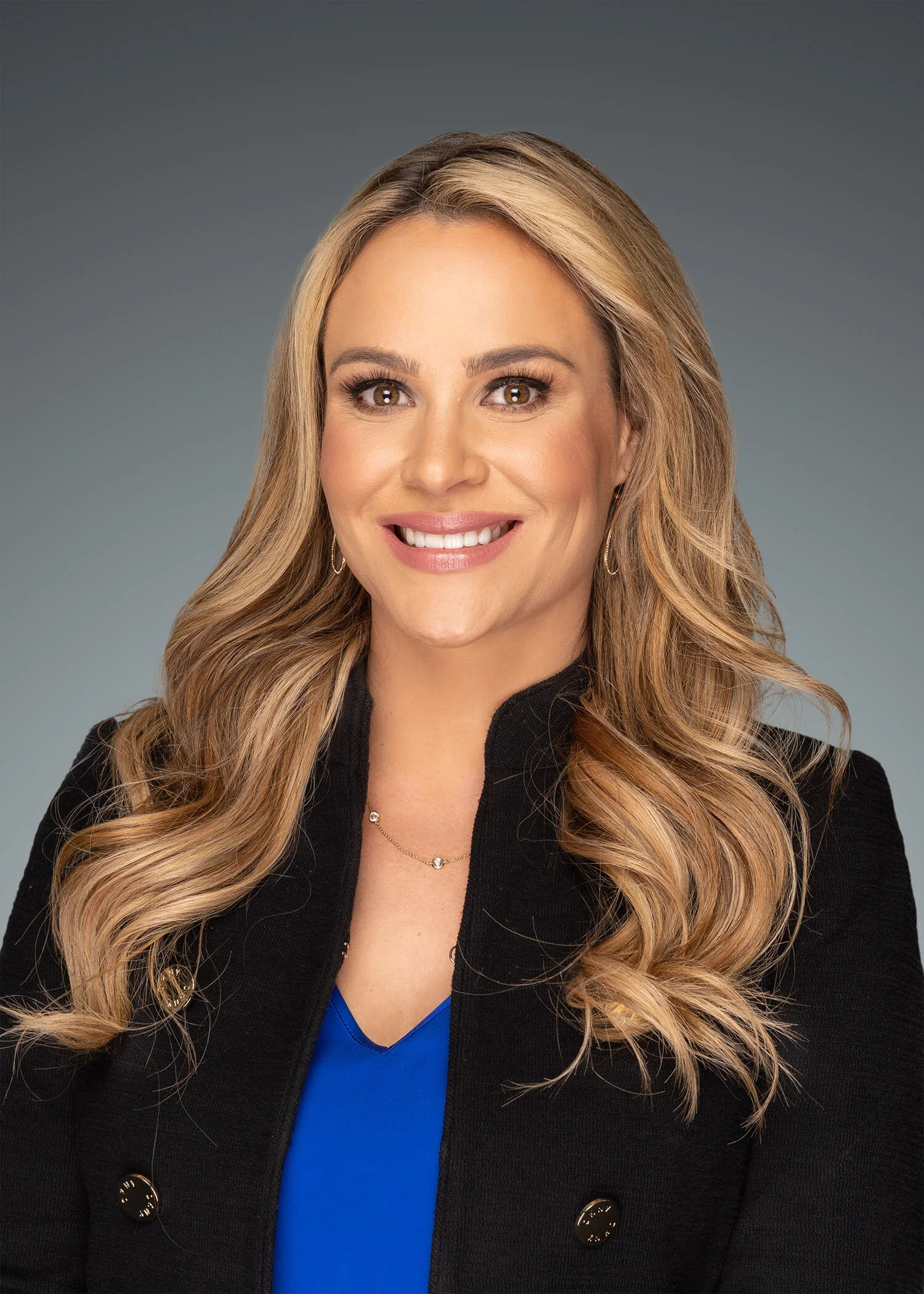 A woman with long, wavy blonde hair, wearing a black jacket with gold buttons, a blue top, and jewelry, smiling at the camera against a gray background.