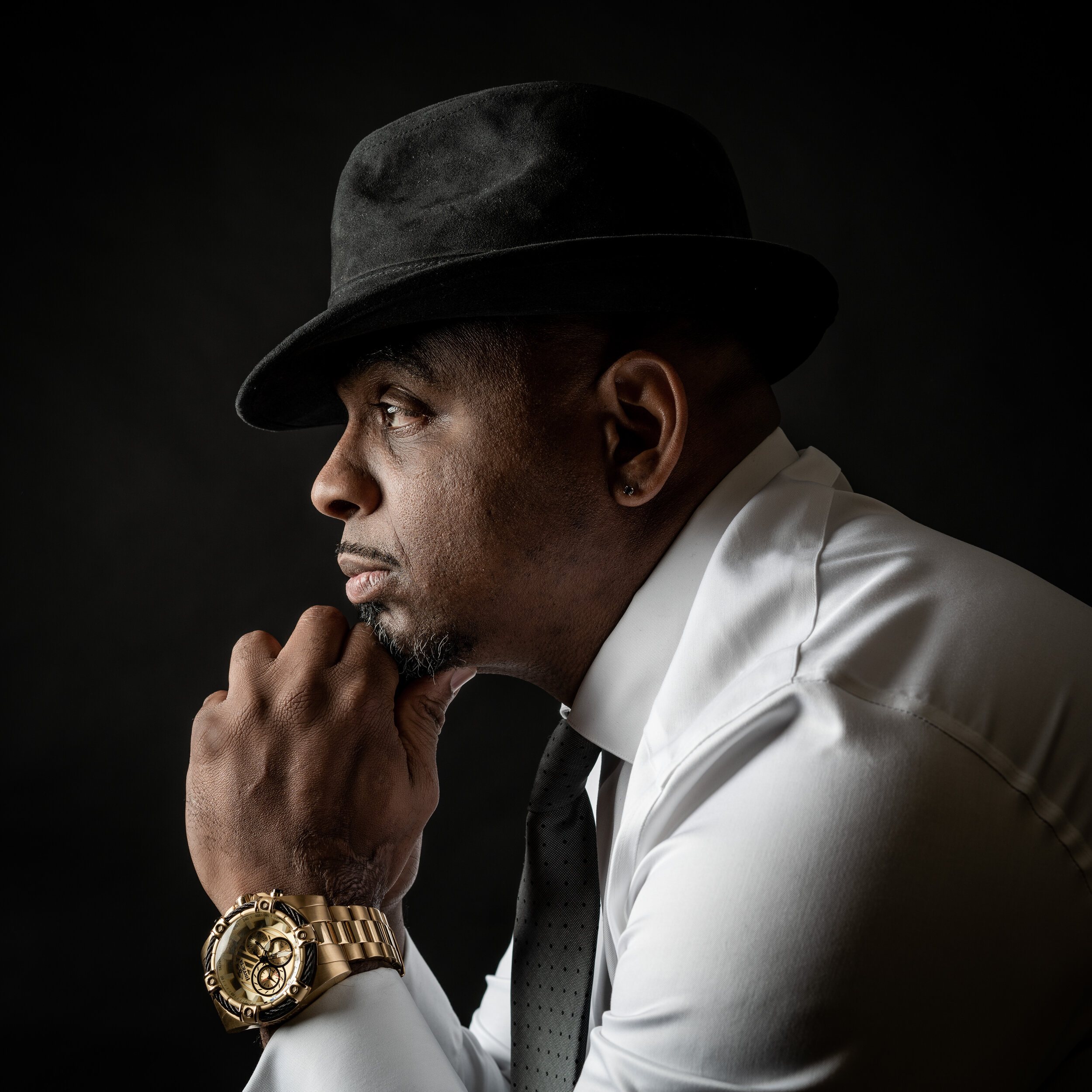 Side profile of a man in a black fedora, white shirt, black tie, with a gold watch, against a dark background.