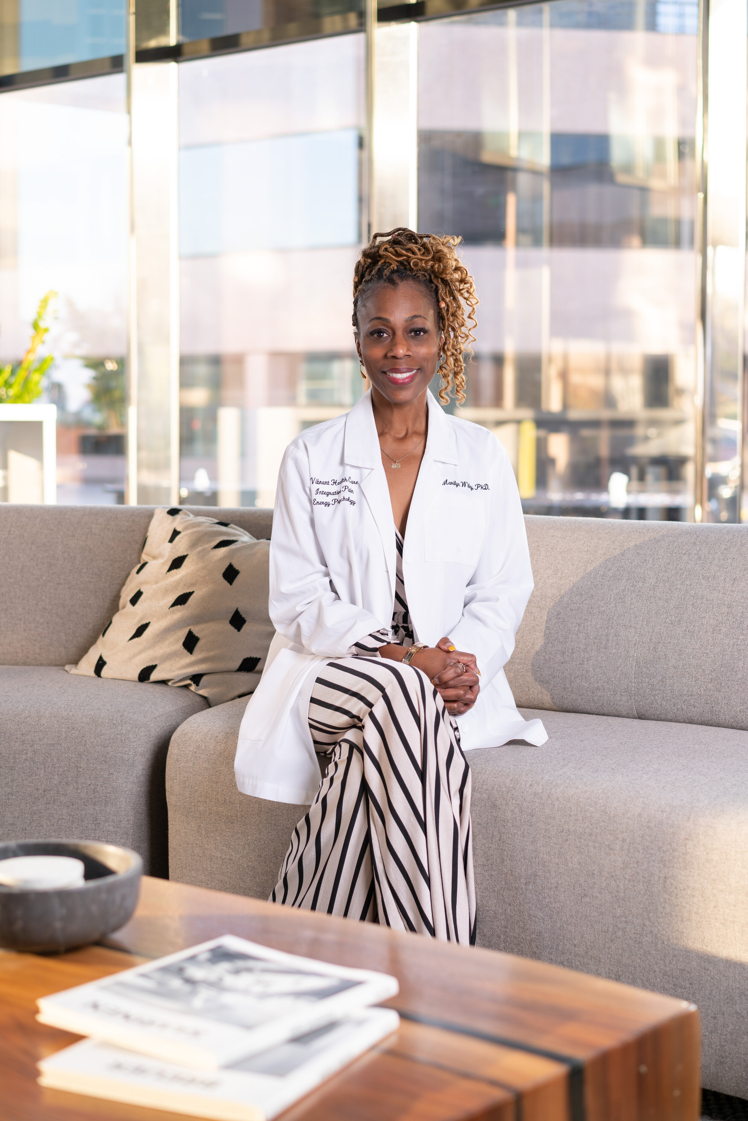 A woman sits on a beige couch in a modern office space with large windows behind her. She is wearing a white coat and striped pants, smiling at the camera.