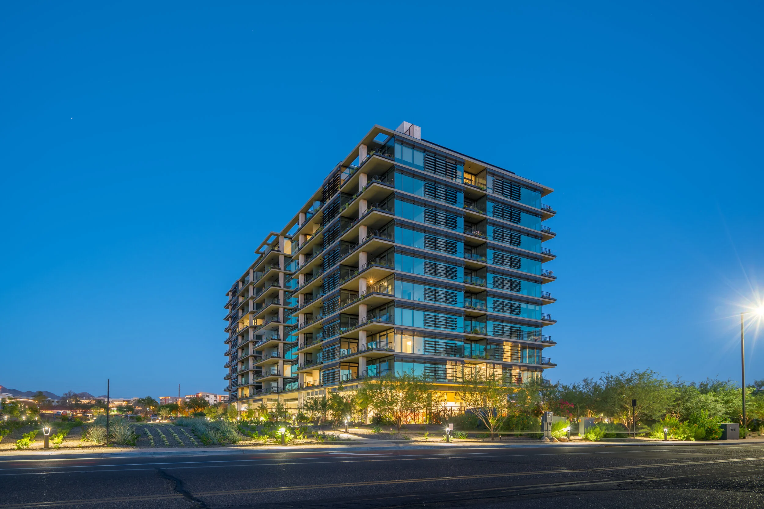 Modern multi-story residential building with glass balconies, illuminated at dusk, with landscaped front yard and street lights.