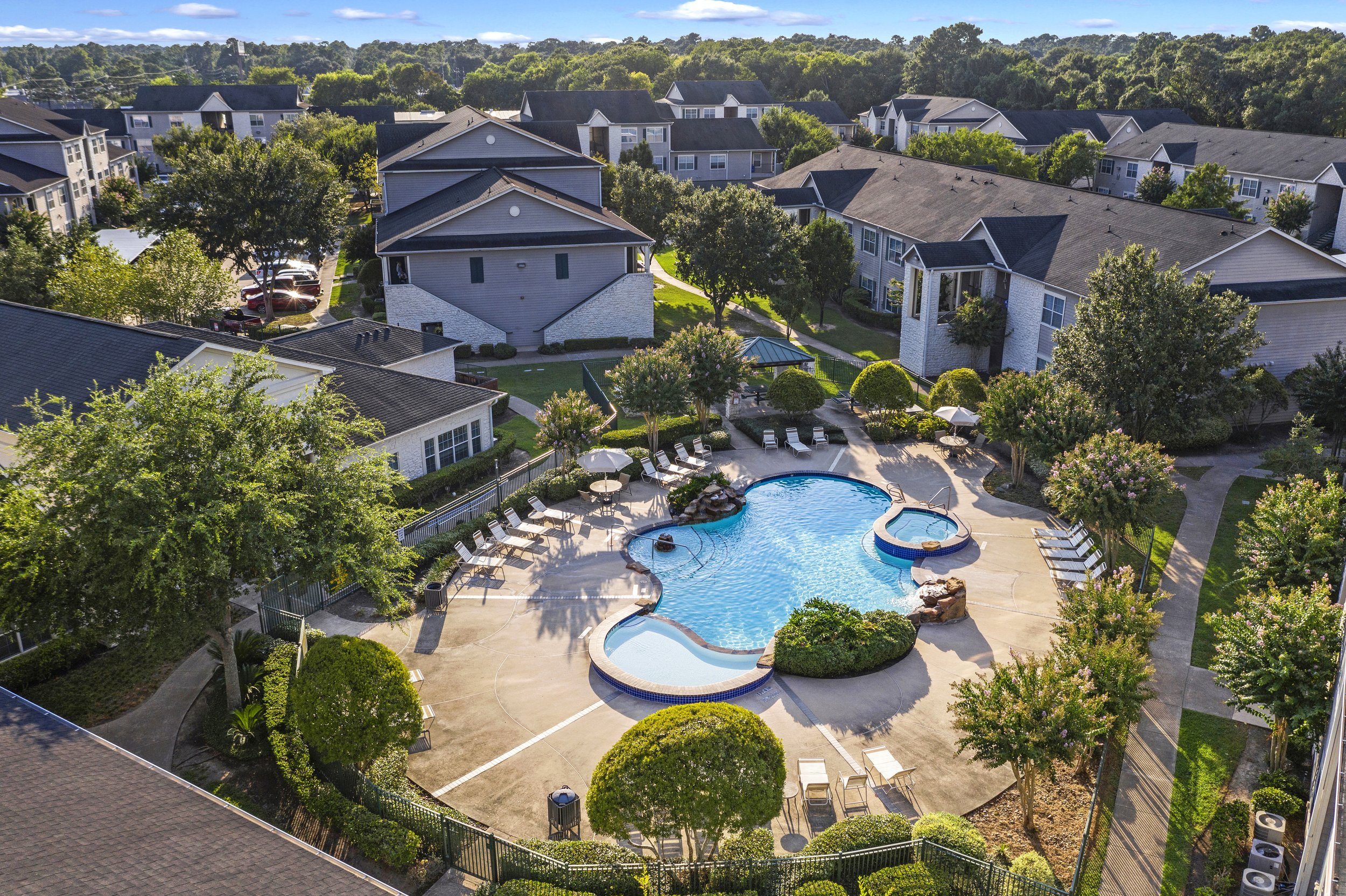 Aerial view of a pool area in an apartment complex, featuring a large freeform swimming pool with a hot tub, surrounded by lounge chairs, umbrellas, trees, and landscaping, with residential buildings and green trees in the background.