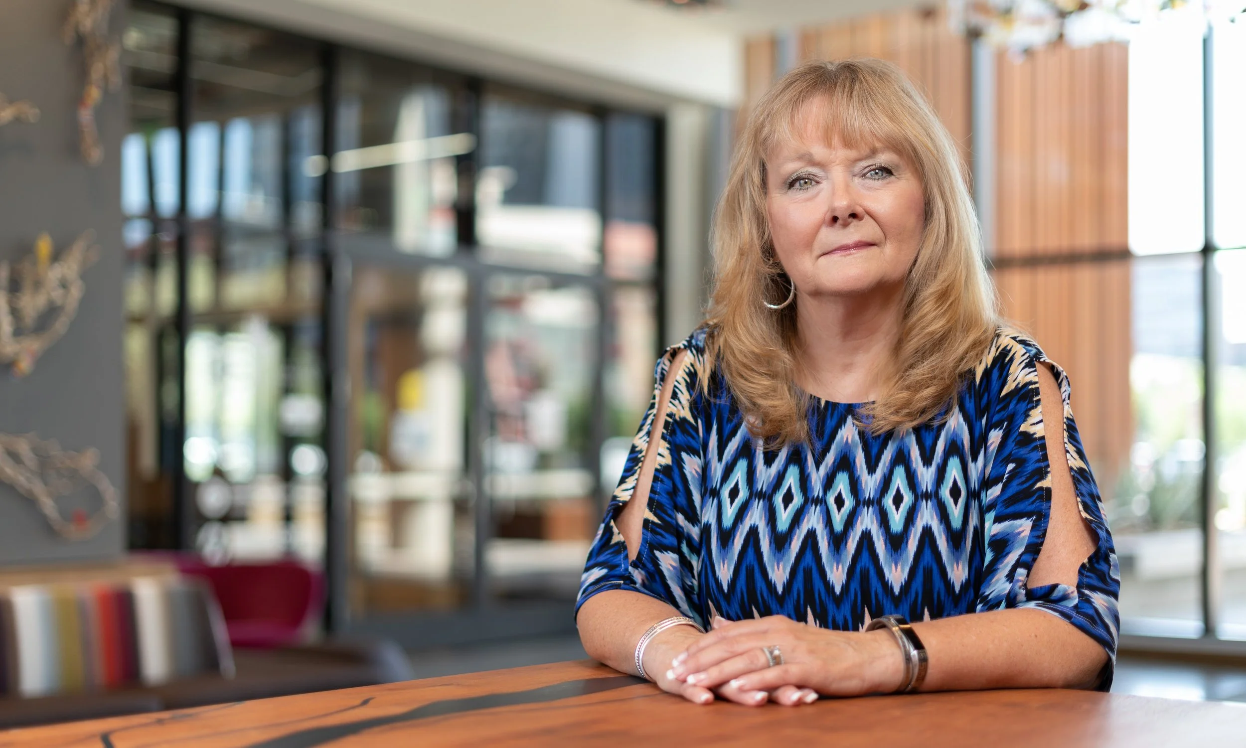 A woman with blonde hair sitting at a wooden table inside a modern cafe or restaurant with large windows and decorative wall art.