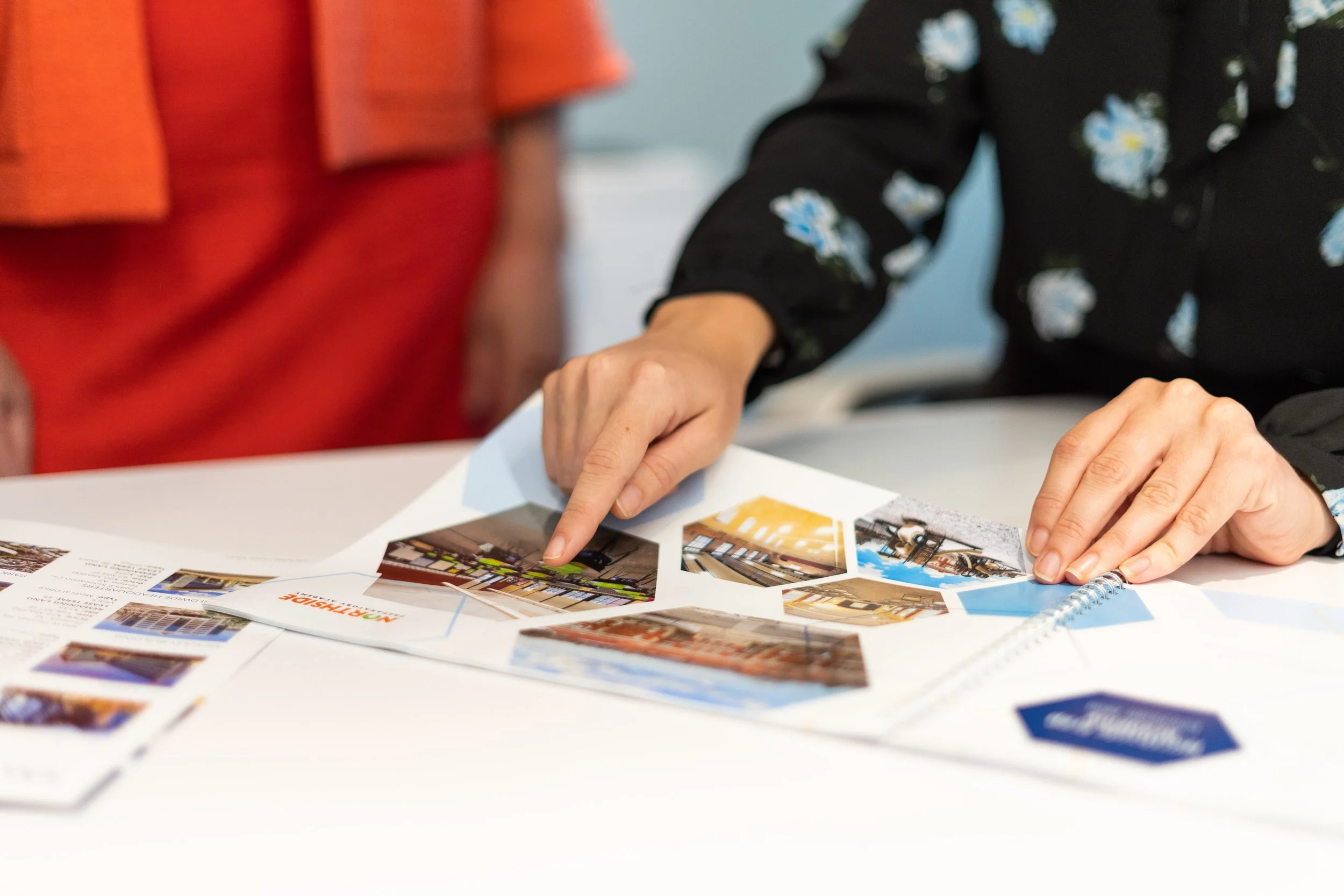 Two people reviewing travel brochures and photographs on a table, with one person pointing at a photo.