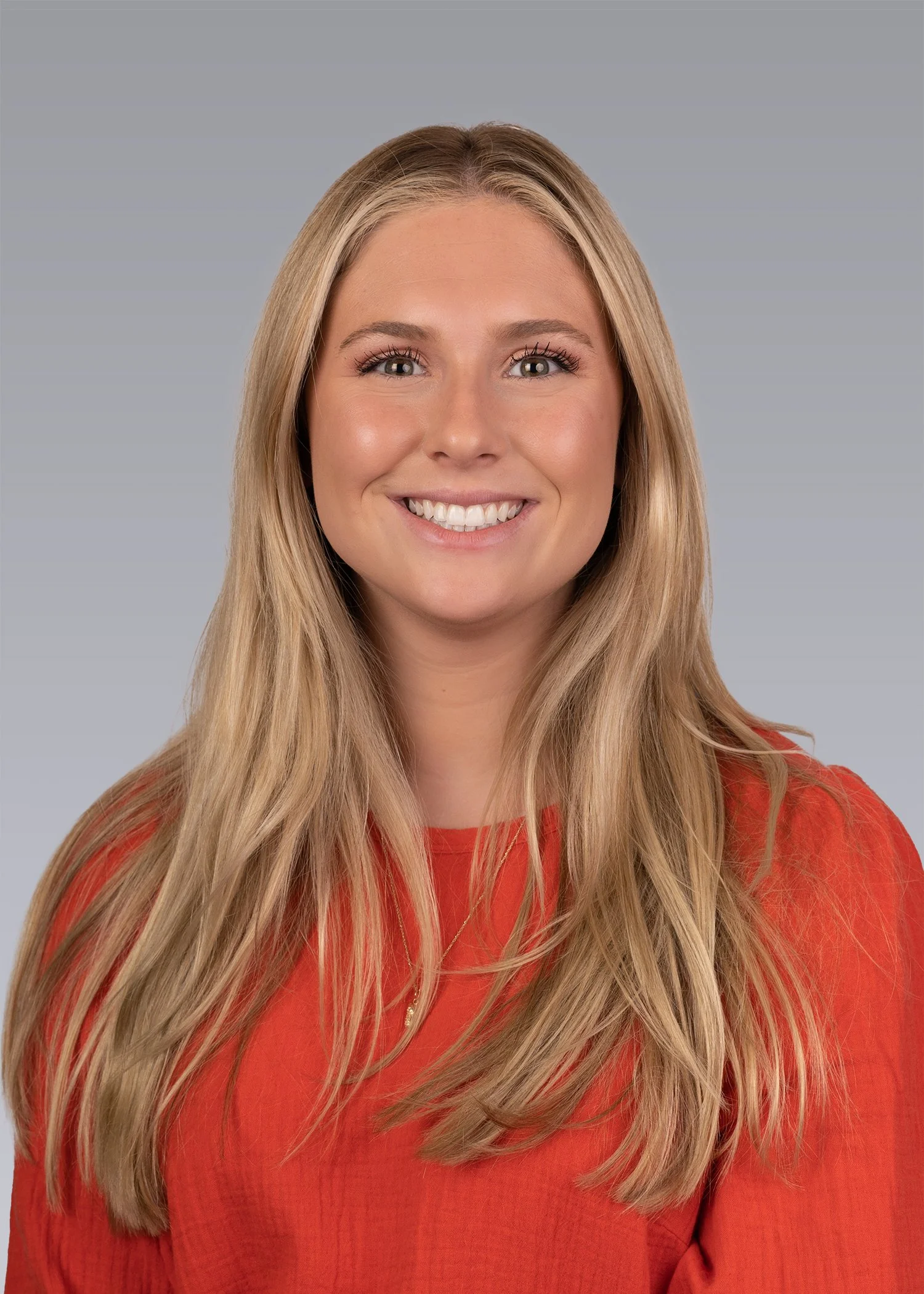 Portrait of a woman with long, blonde hair, smiling and wearing a red top, against a plain gray background.