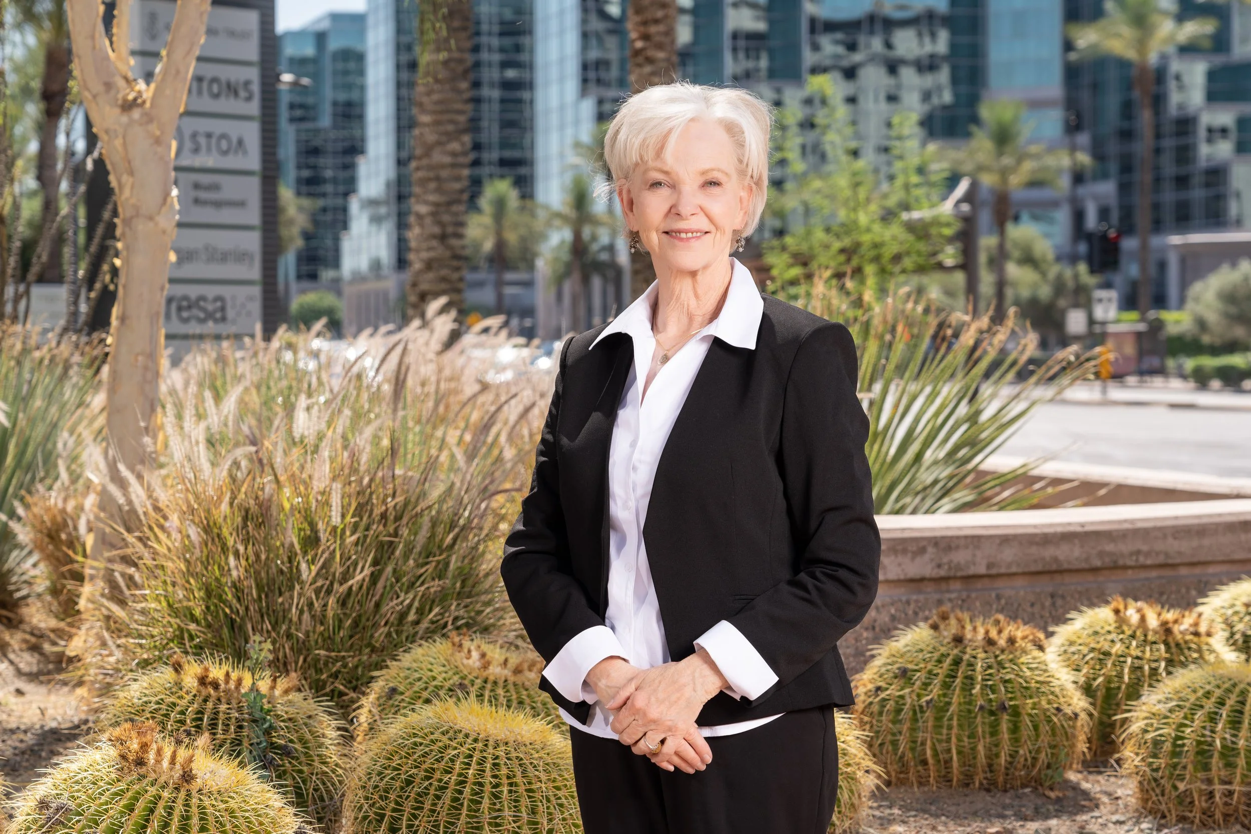 An elderly woman with short gray hair wearing a black blazer and white shirt, smiling outdoors near desert plants and cacti, with modern buildings in the background.