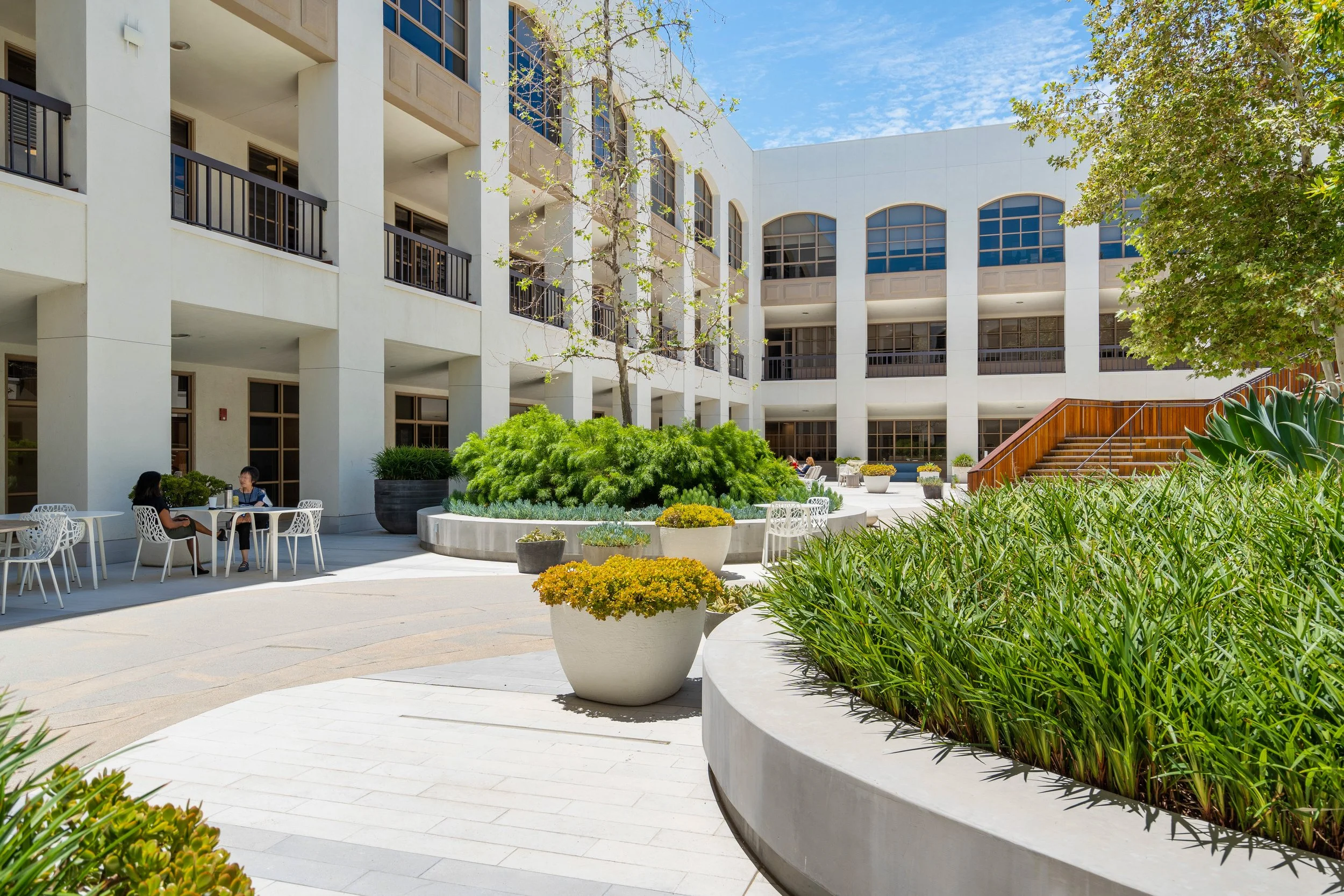 An outdoor courtyard in front of a modern, multi-story building with large windows. The courtyard features various plants in large pots, green shrubs, and trees, with a few people sitting at white tables and chairs.