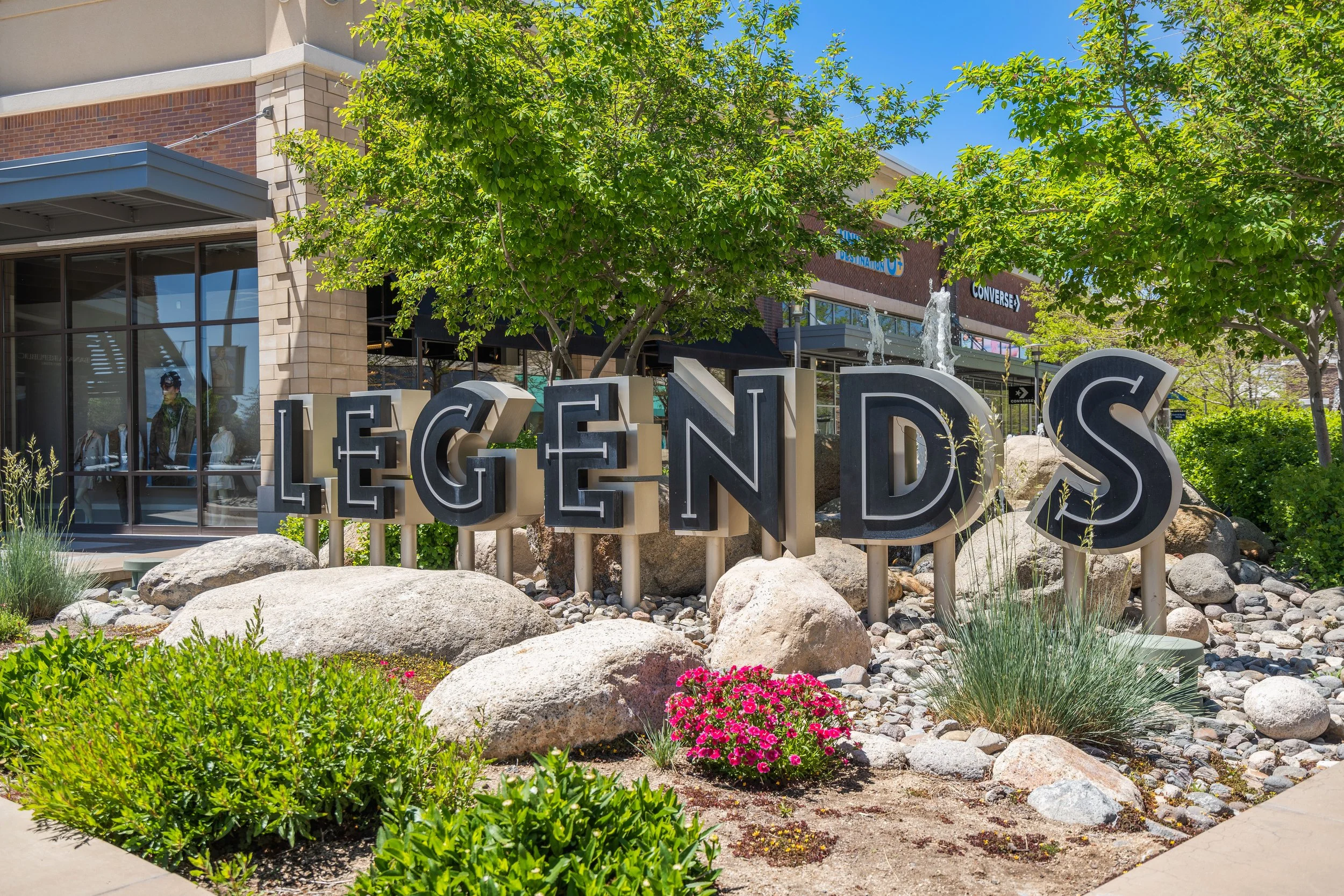 Sign spelling 'LEGENDS' in front of a landscaped area with rocks, plants, and trees, with stores and fountains in the background.