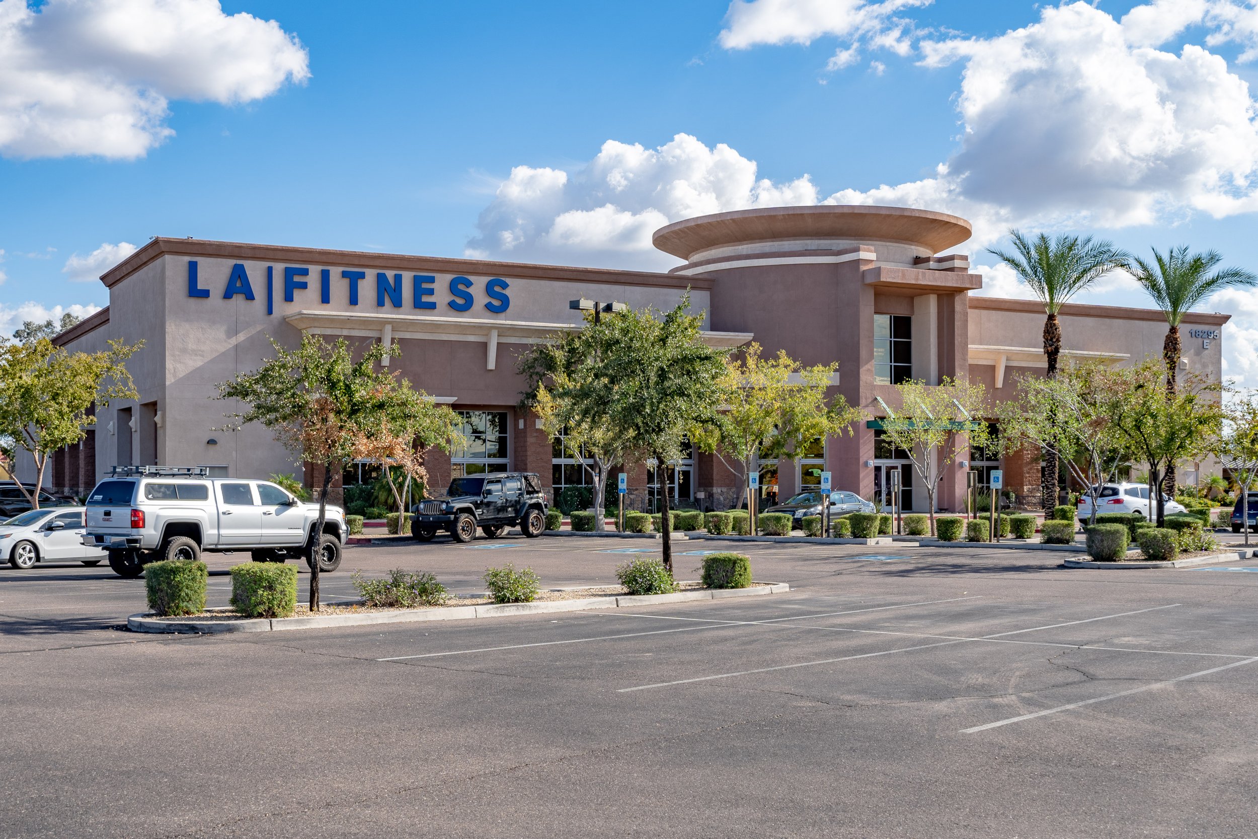 Front view of a fitness center building with a large blue sign saying 'LA FITNESS', surrounded by a parking lot with several parked cars, small trees, and palm trees under a partly cloudy sky.