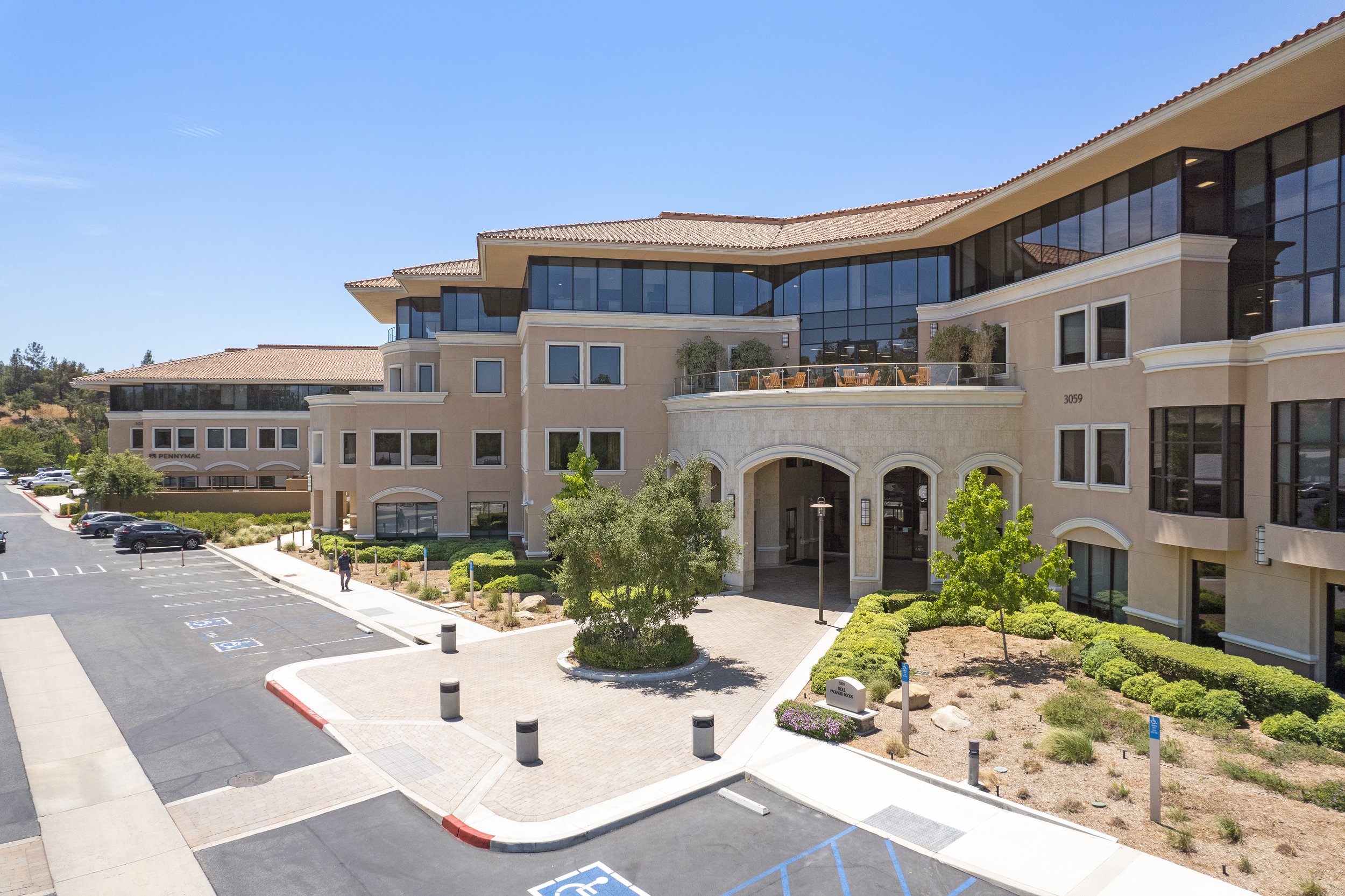 Modern multi-story office or residential building with large glass windows, surrounded by a landscaped parking lot with designated handicapped parking spots, trees, and bushes on a bright, sunny day.