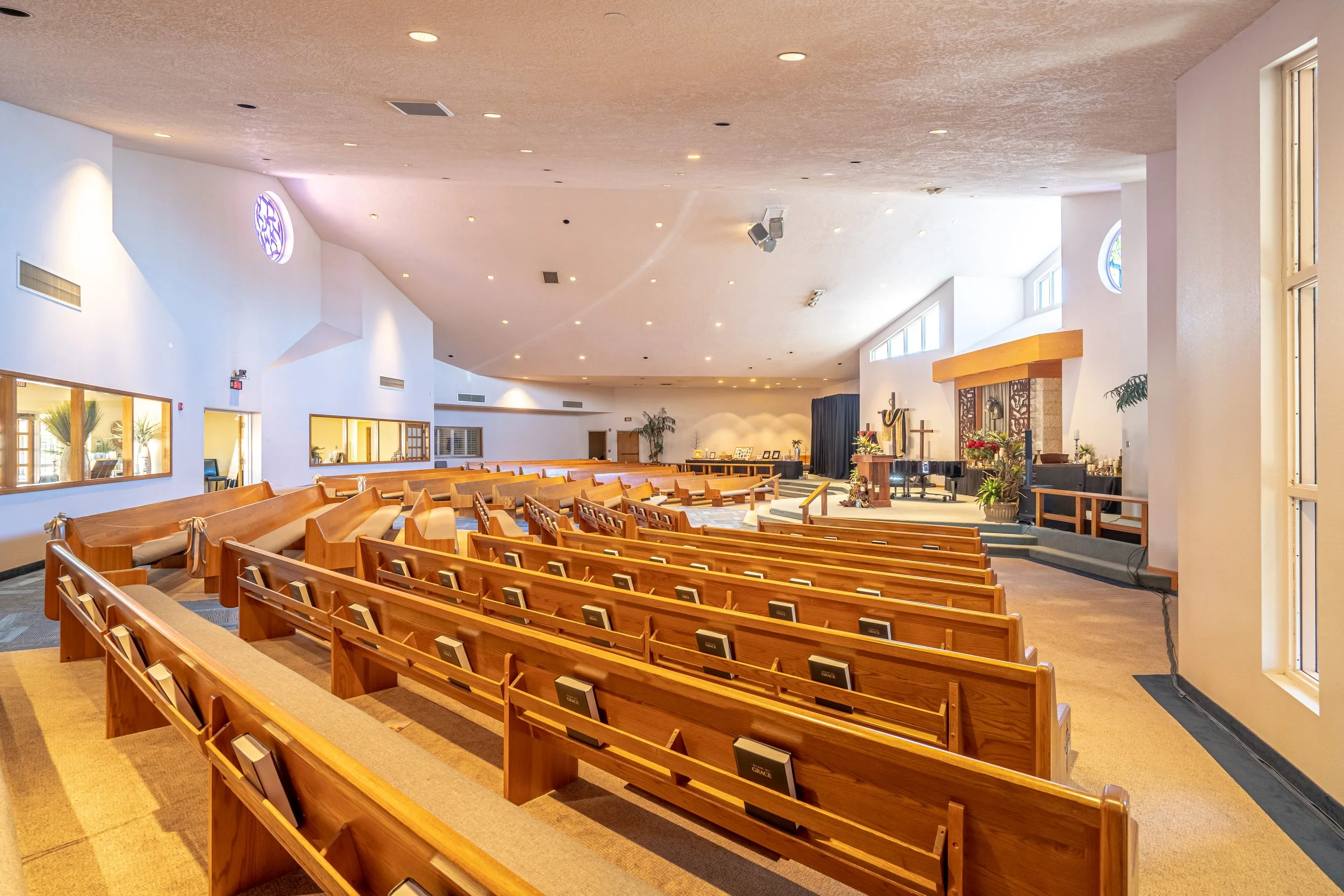 Empty church sanctuary with wooden pews facing an elevated stage with a piano, cross, and religious decorations.