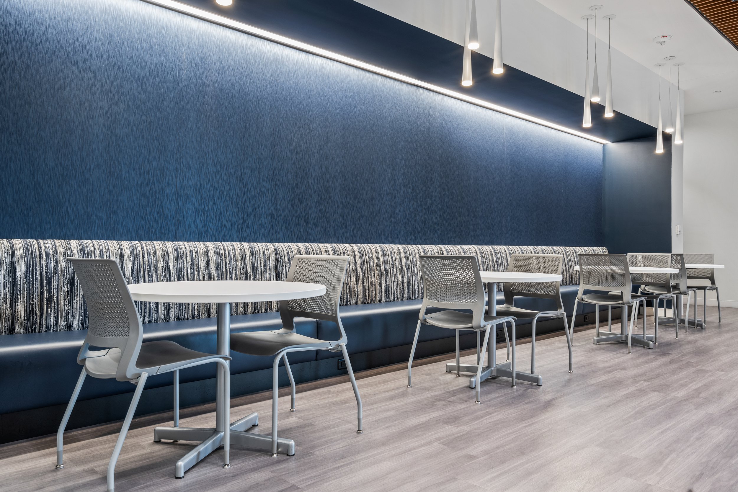 Modern interior with white tables, gray chairs, a long blue patterned bench along a blue textured wall, and contemporary pendant lighting.