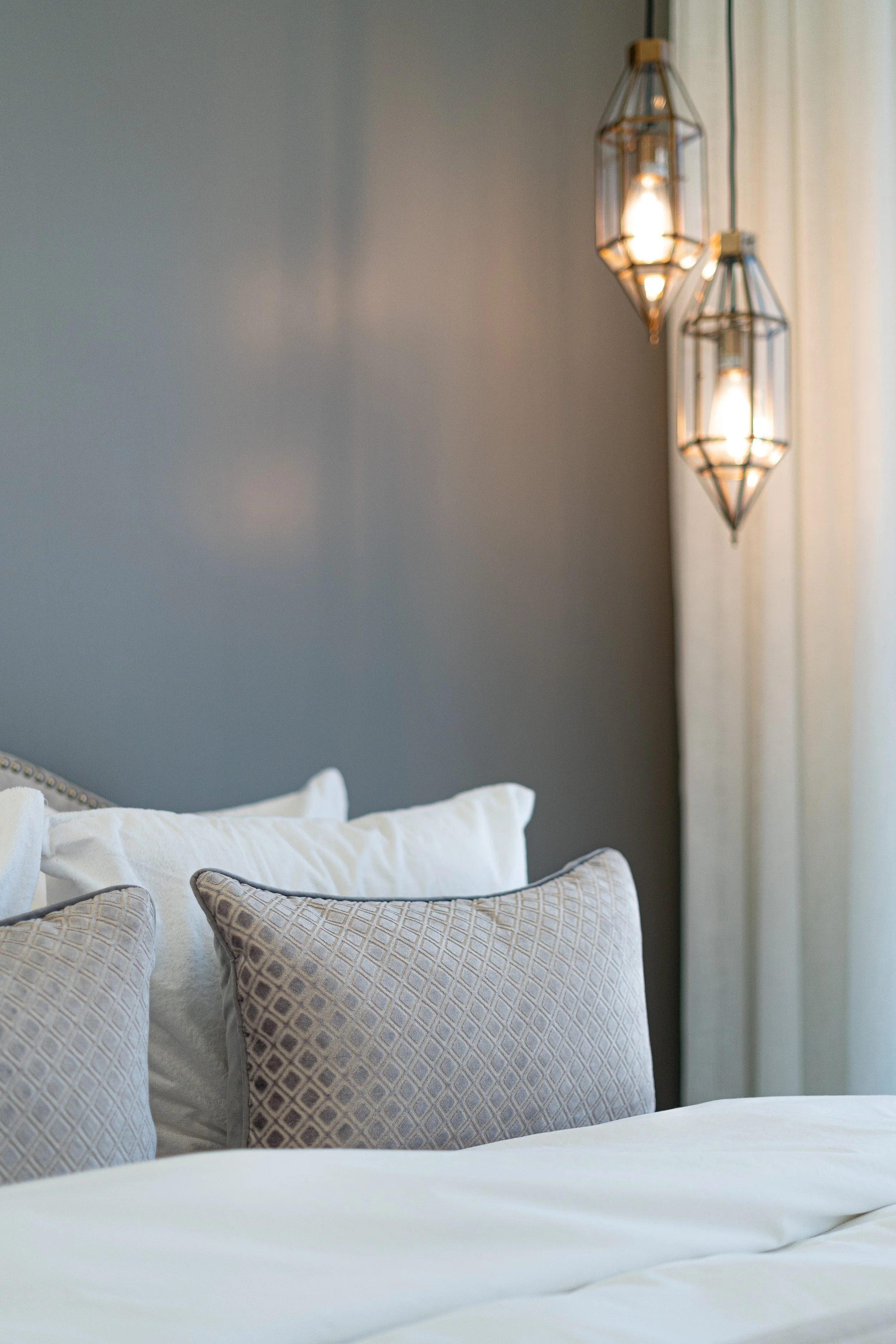 A cozy bedroom with a bed adorned with white and patterned pillows, gray wall, hanging pendant lights, and sheer curtains by a window.