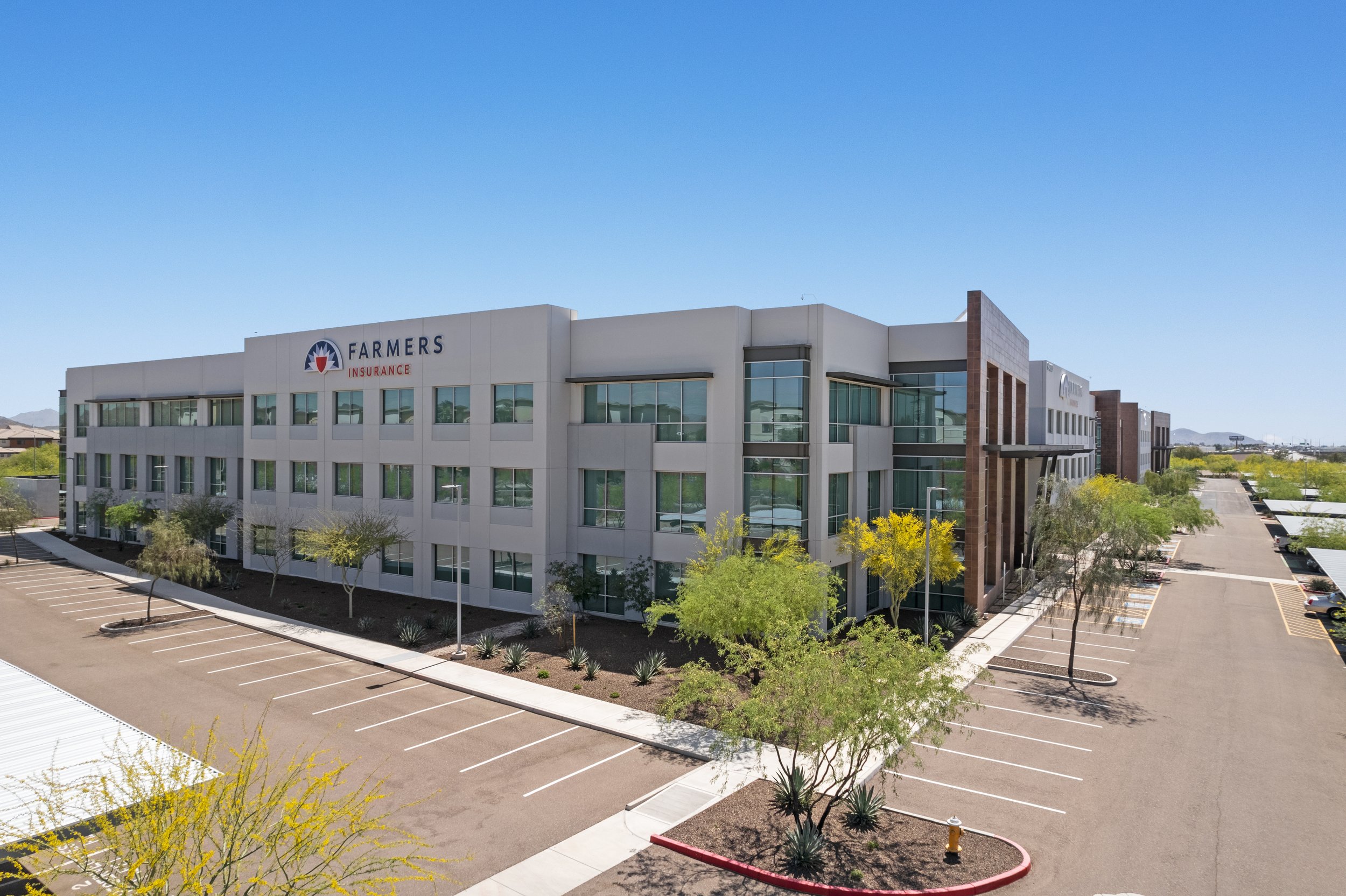Exterior view of a modern office building with a sign that reads Farmers Insurance, surrounded by parking lots and landscaped with trees and bushes, under a clear blue sky.