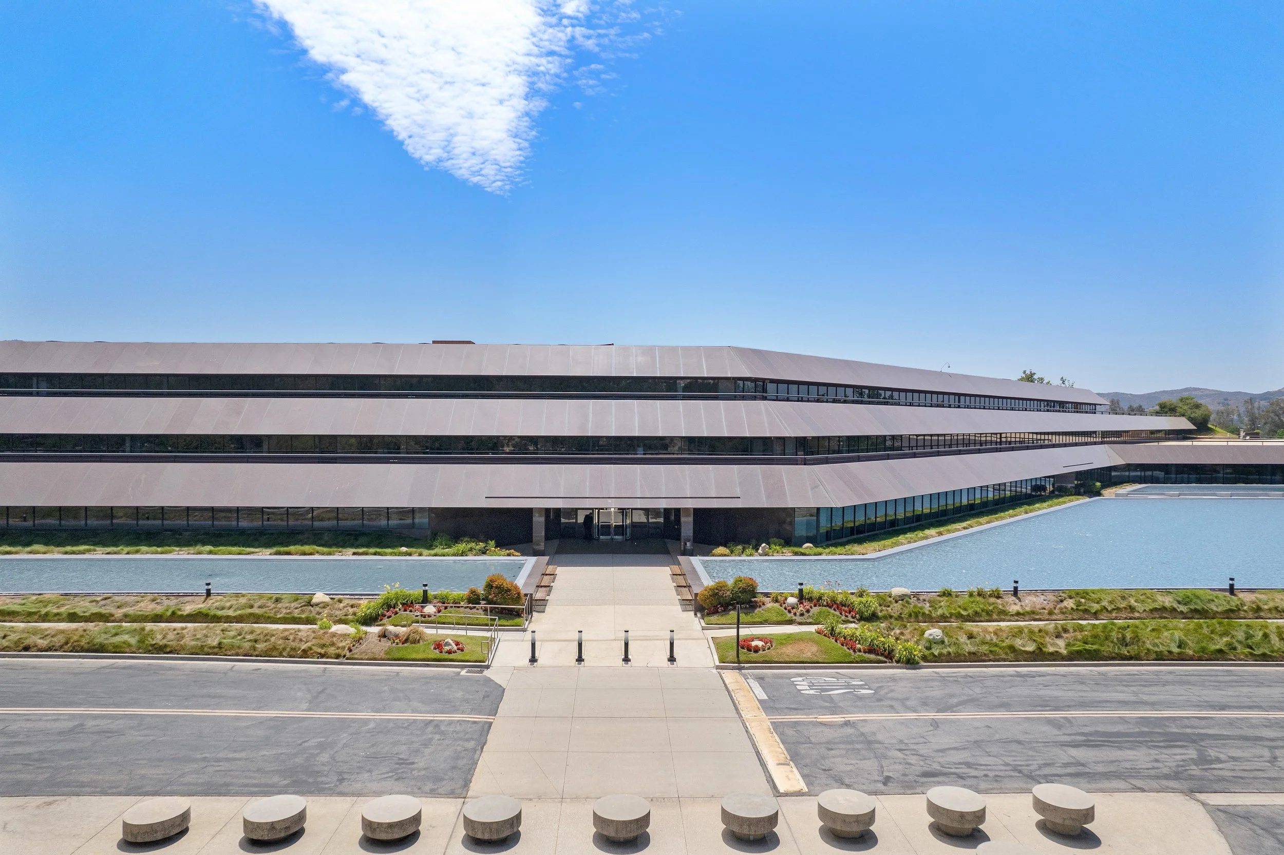 Modern multi-story building with horizontal lines, surrounded by water features, landscaped gardens, and a clear blue sky.
