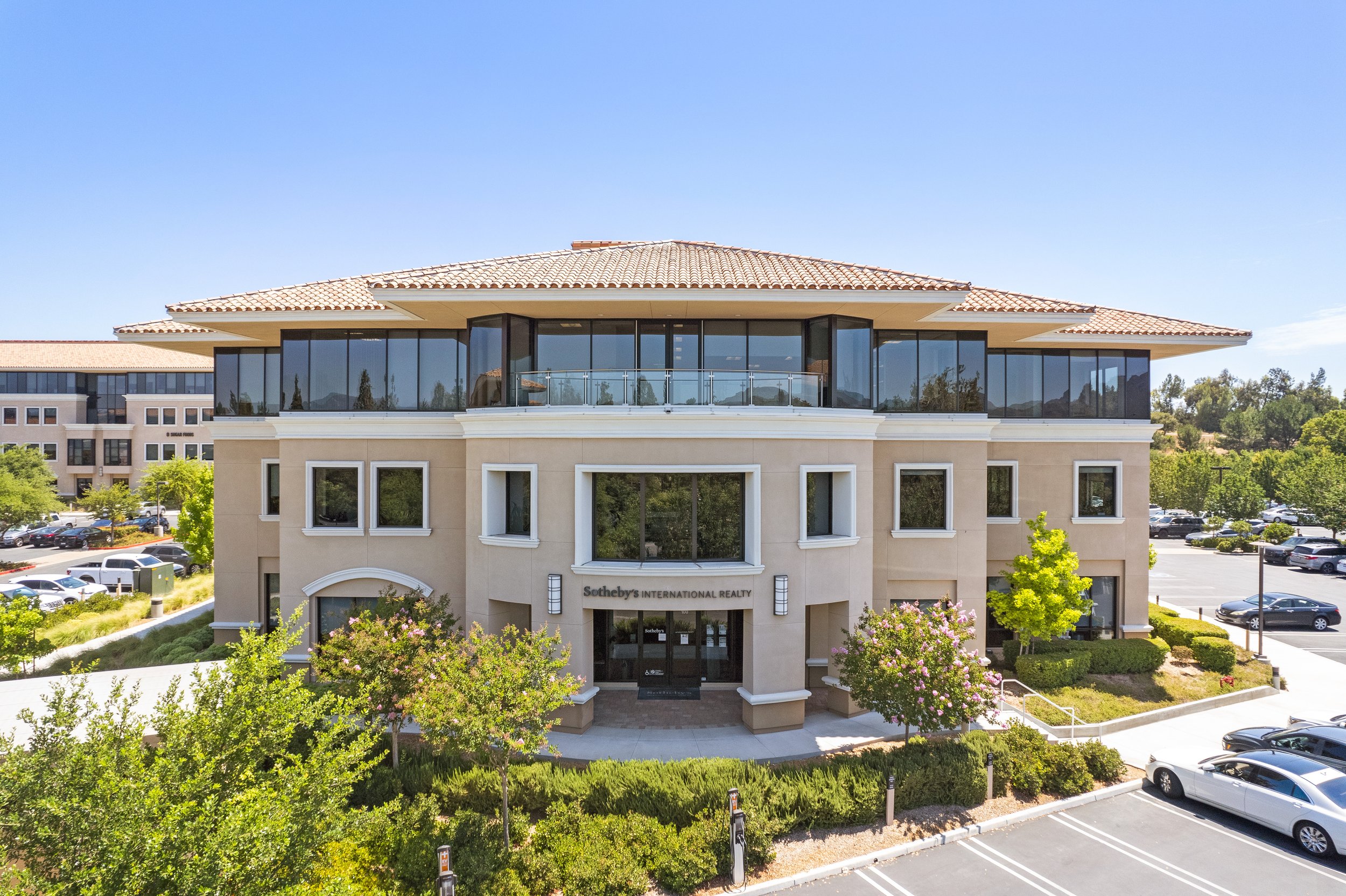 Exterior view of a modern commercial building with beige walls, large windows, and a sign for Sotheby's International Realty, surrounded by parking lots, trees, and landscaped greenery.