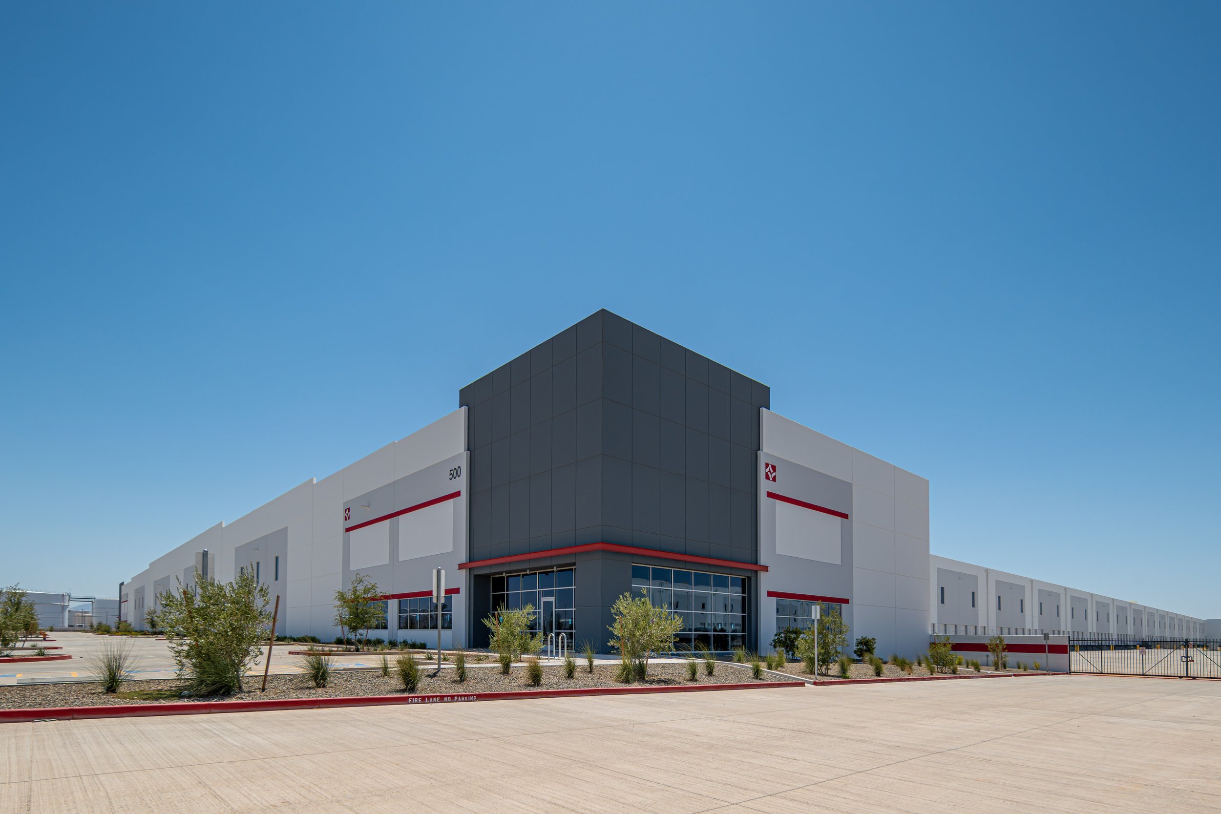 Modern warehouse building with white exterior, black accents, and a red stripe, surrounded by a paved lot and sparse desert landscaping under a clear blue sky.
