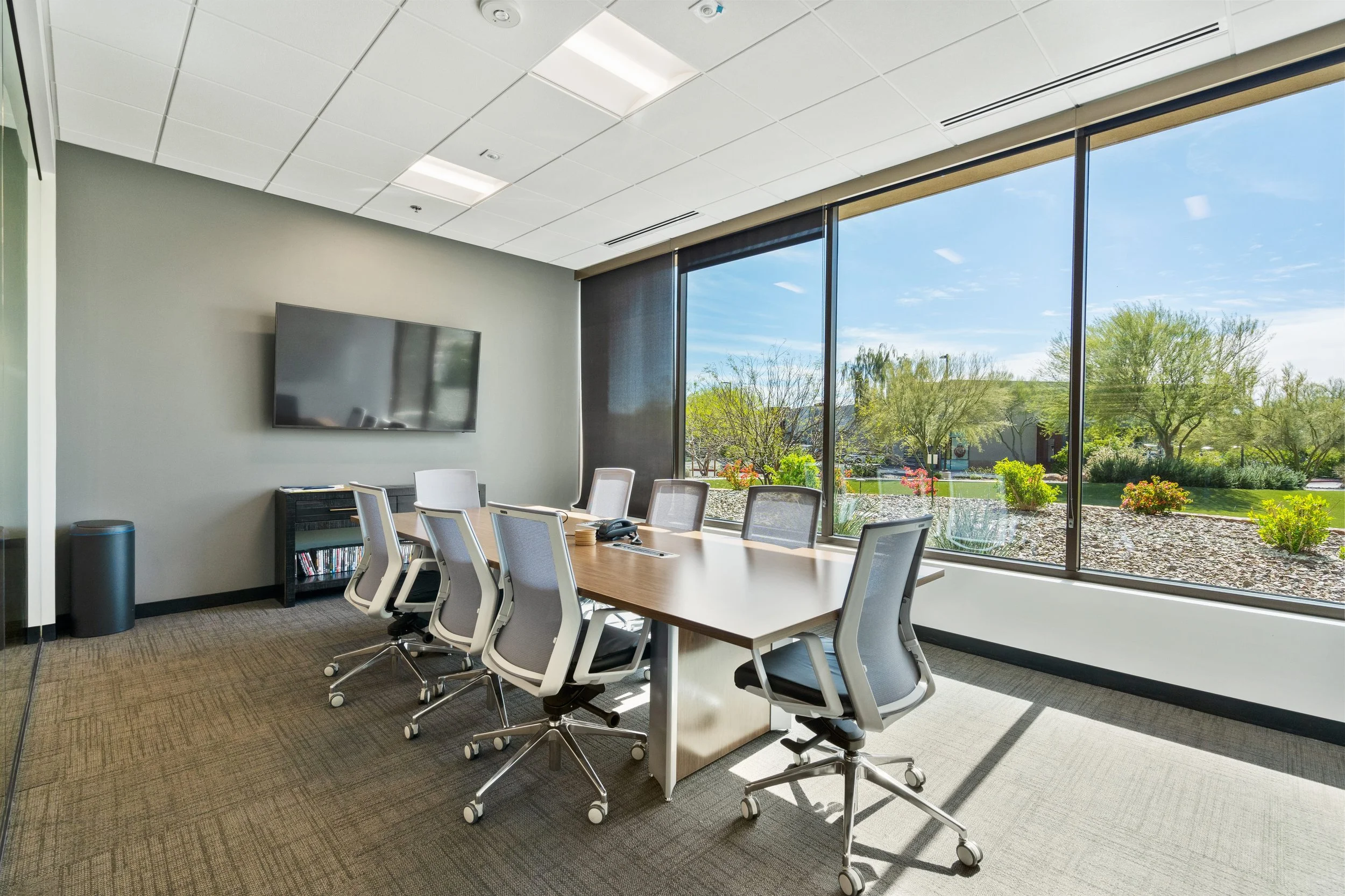 Modern conference room with large window showing outdoor landscaping, six white office chairs around a rectangular wooden table, wall-mounted TV, small black media stand, and a trash bin.