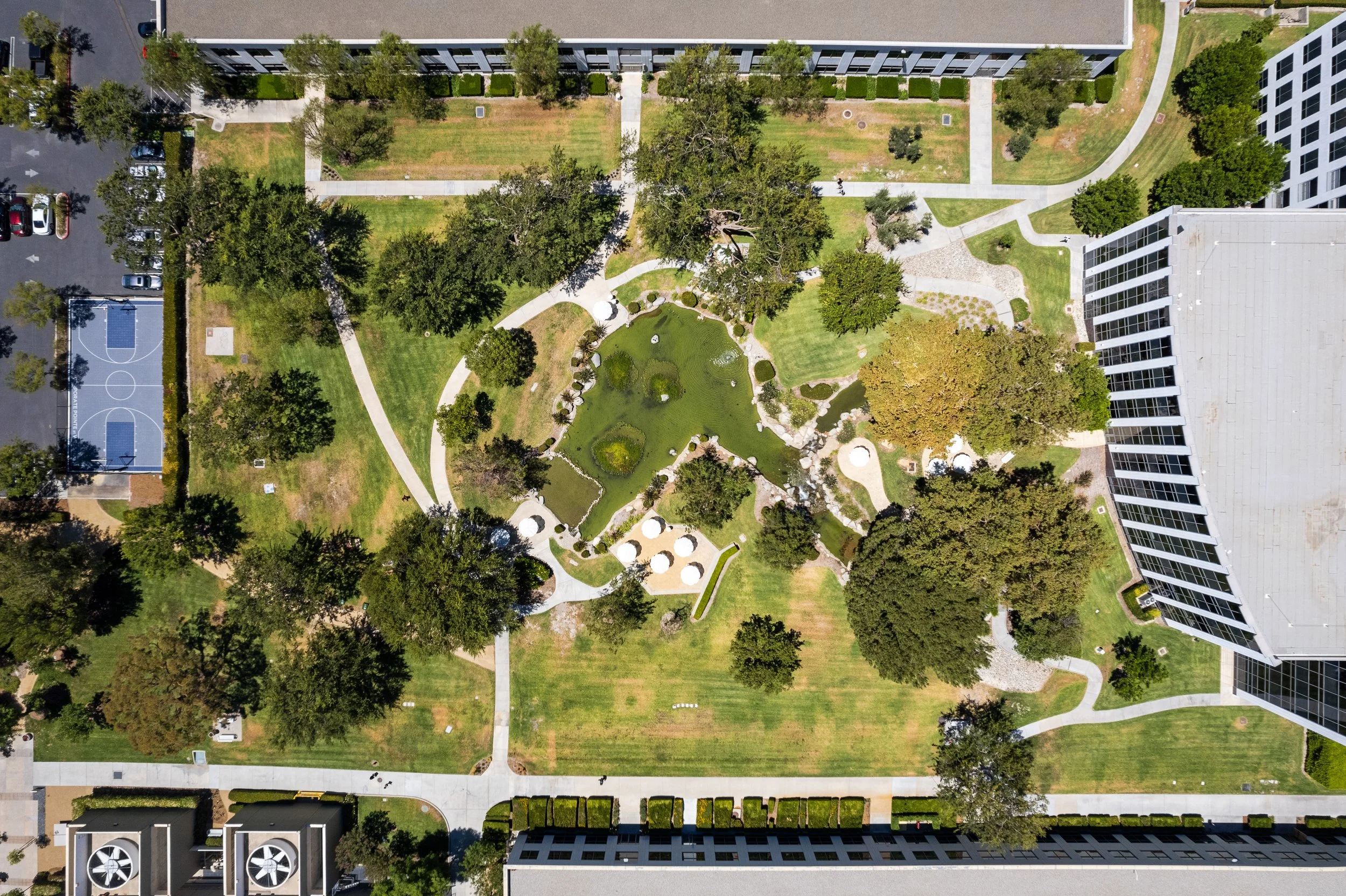 An aerial view of a landscaped courtyard with pathways, trees, grass, and a small pond, surrounded by multi-story residential buildings and a basketball court.