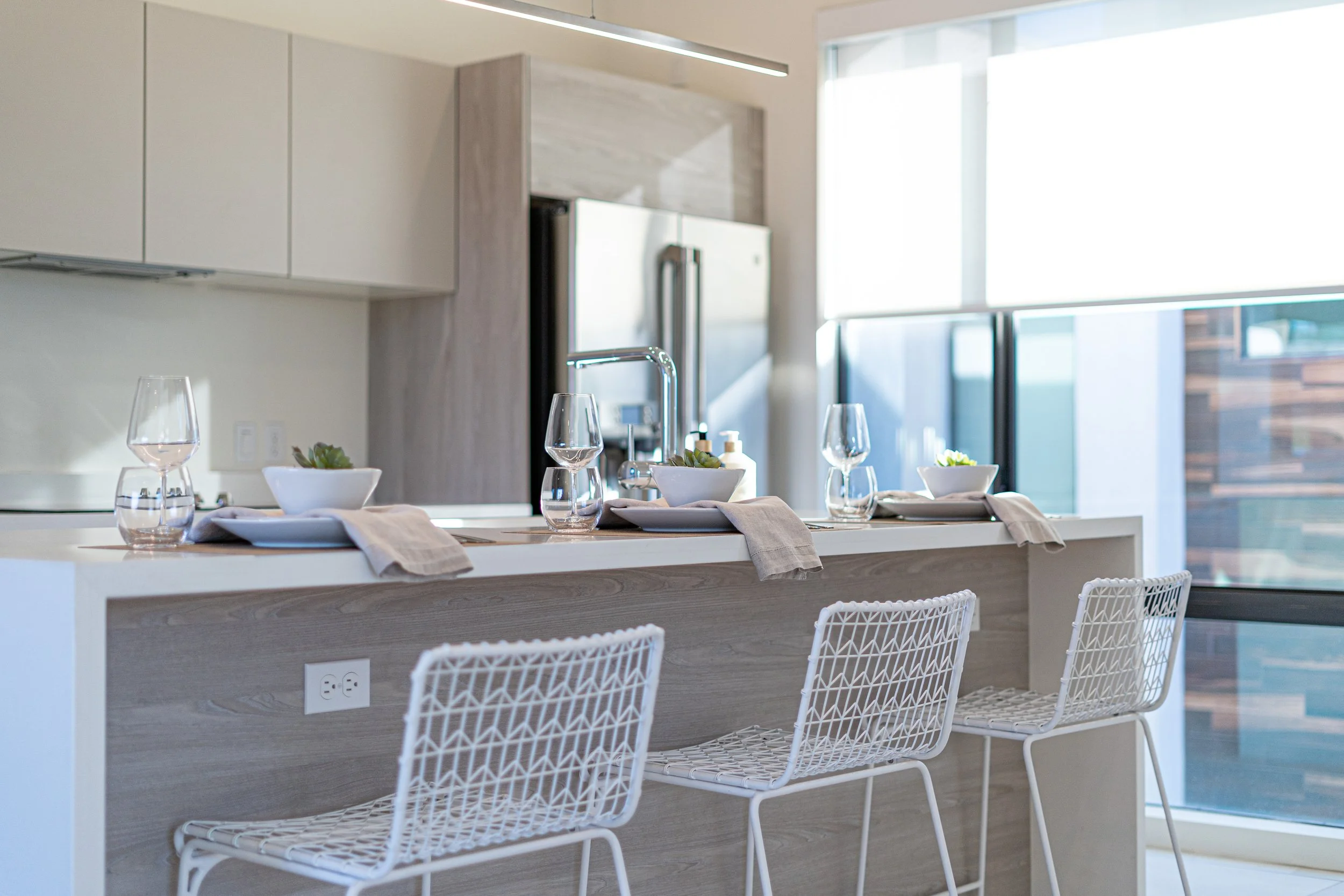 Modern kitchen with a breakfast bar set for three, three white wire chairs, white cabinets, a stainless steel refrigerator, and large windows with a view of neighboring buildings.