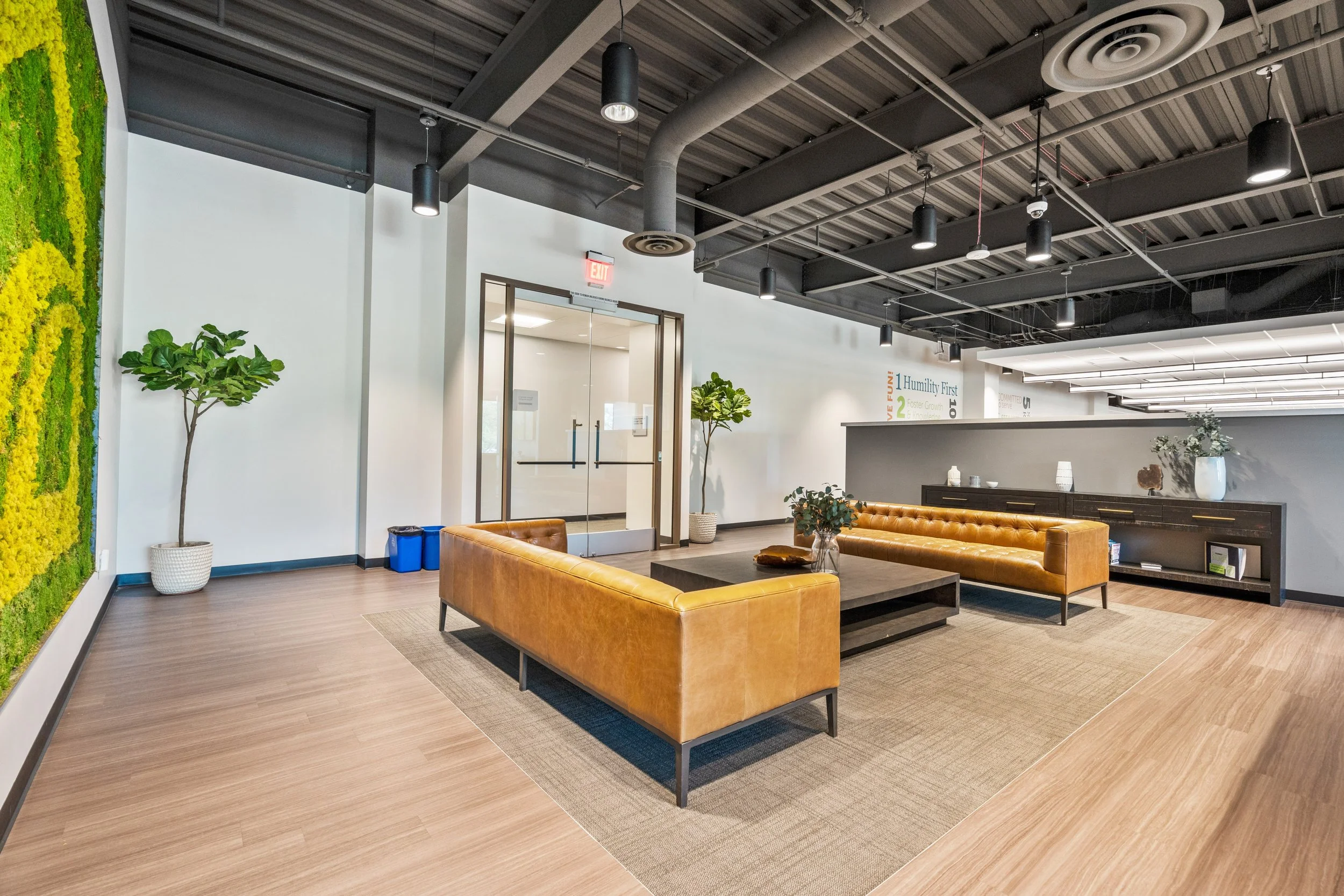 Modern office lobby with two brown leather couches surrounding a black coffee table, green plants in pots, a wooden sideboard with decorative items, and a wall with motivational words.