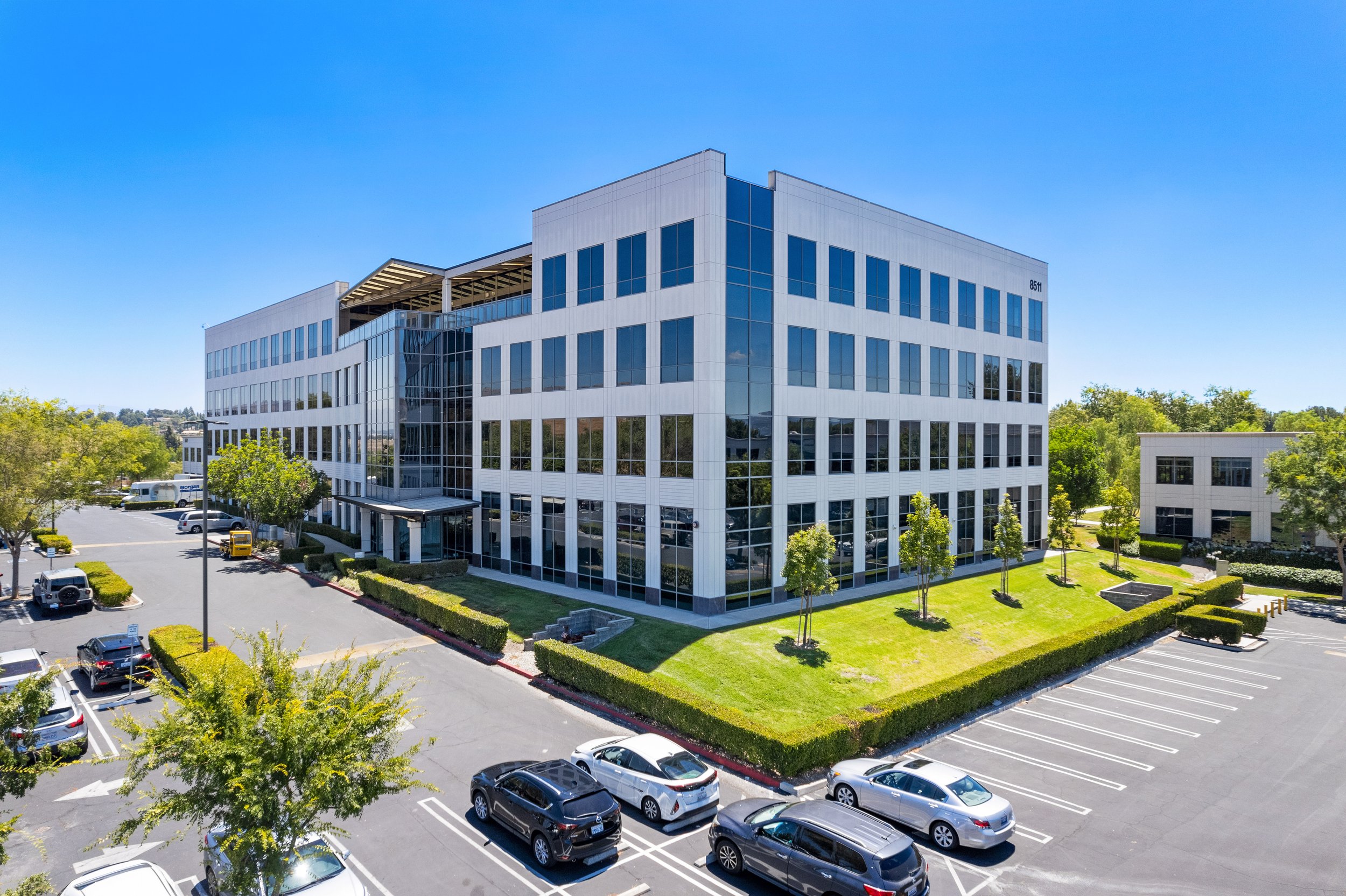 A modern multi-story office building with large glass windows and white exterior panels, surrounded by a parking lot with cars and small trees, under a clear blue sky.