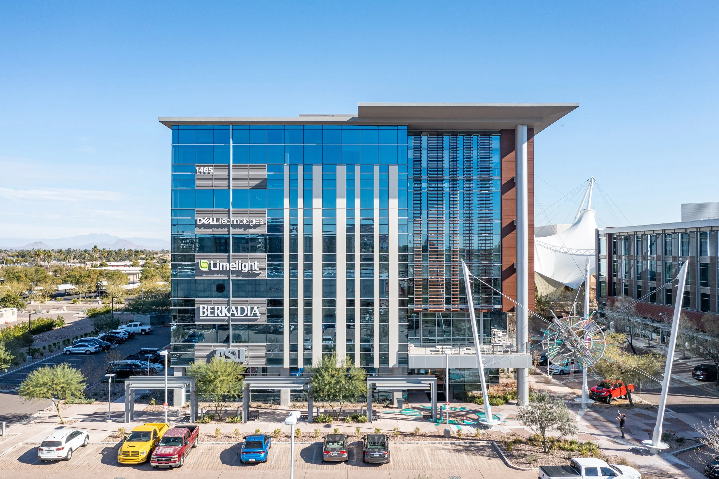 Modern office building with glass facade, displaying company logos such as Dell Technologies, Limelight Networks, Berkadia, and ASU. Several parked cars and trees in the foreground, with a blue sky and distant mountains in the background.