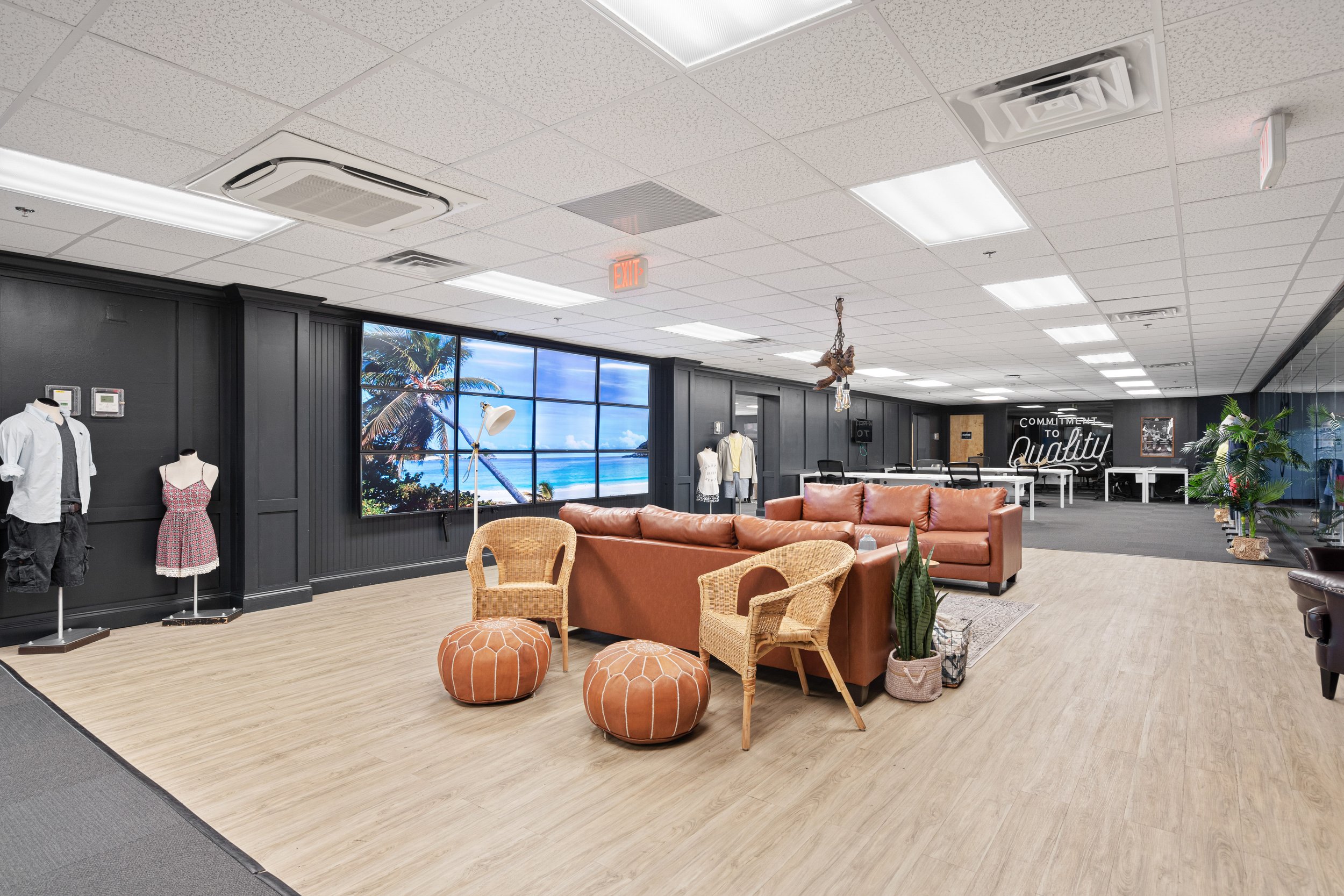 Office lounge area with a large digital wall display showing a beach scene, brown leather sofa, rattan chairs, poufs, indoor plants, and a black wall with white decor text.