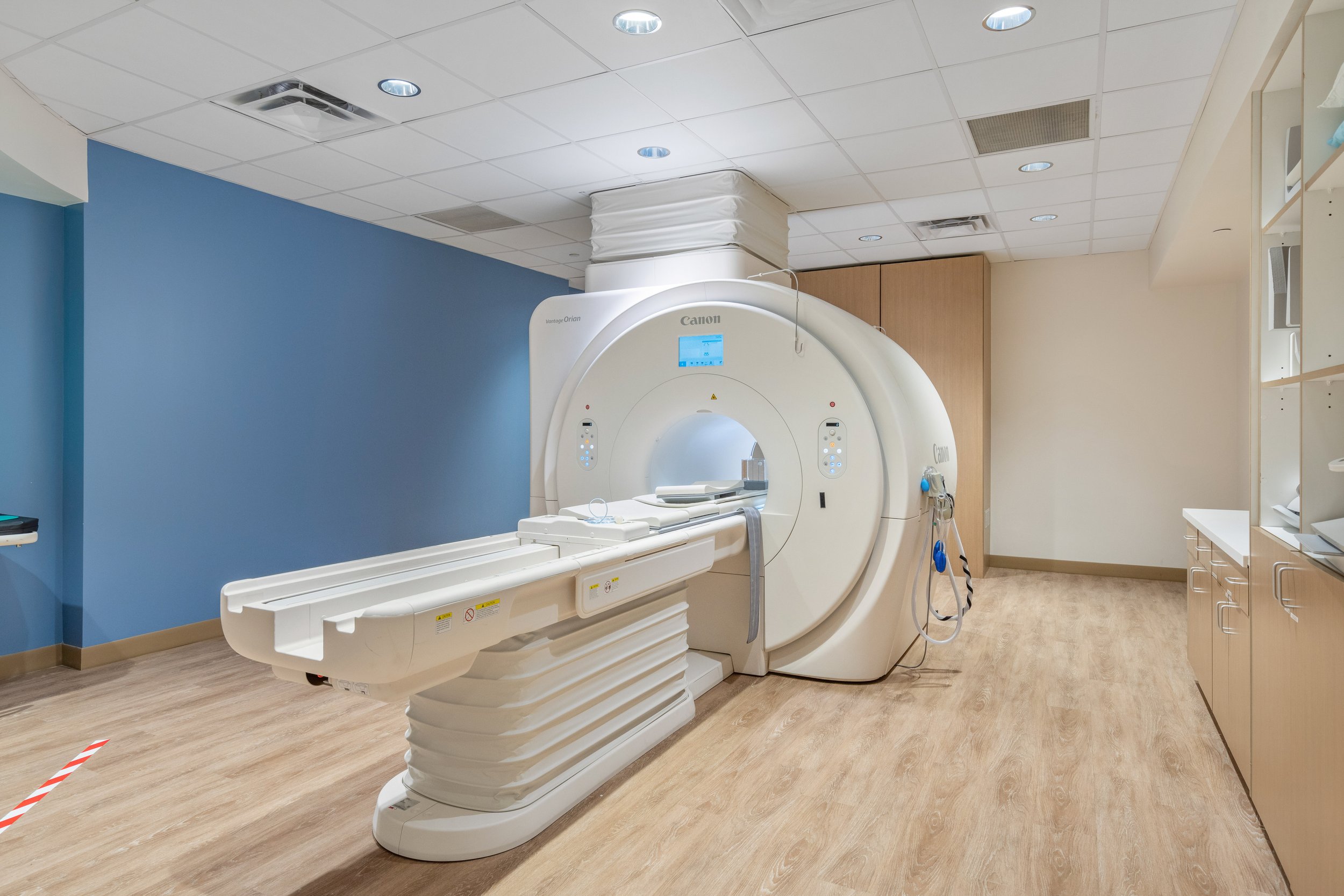 A medical MRI machine in a clean, well-lit room with light wood flooring and light-colored walls.