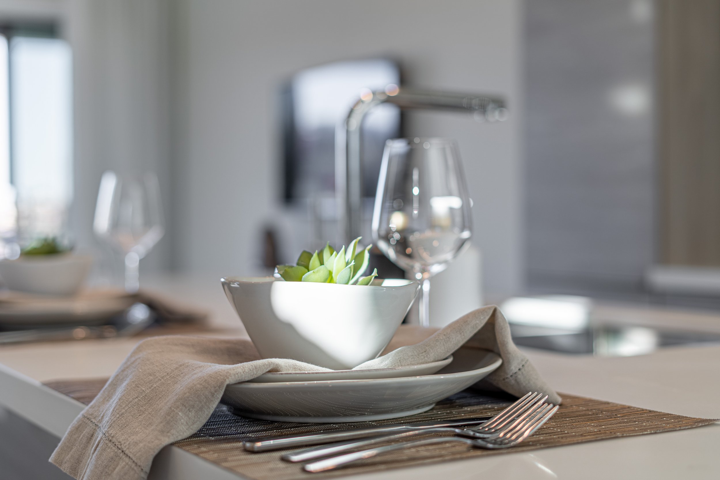 A modern kitchen countertop set for a meal, with a white bowl containing green plants, a white plate, a beige napkin, and silverware, near a water glass and a faucet.