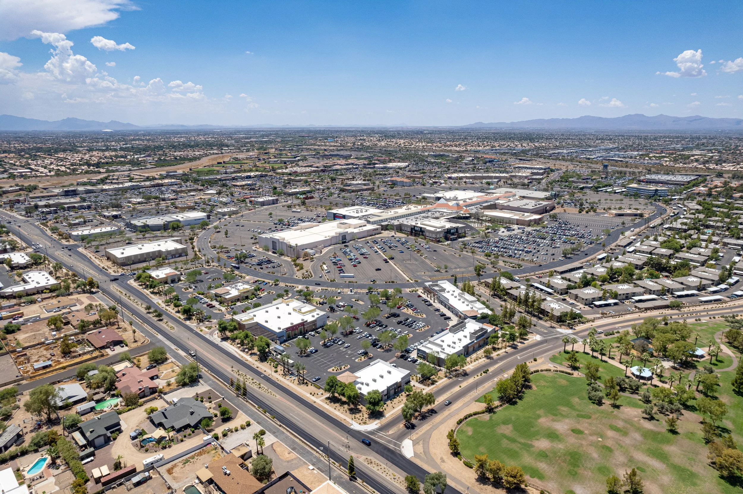 Aerial view of a shopping center and surrounding residential area with mountains in the background under a partly cloudy sky.