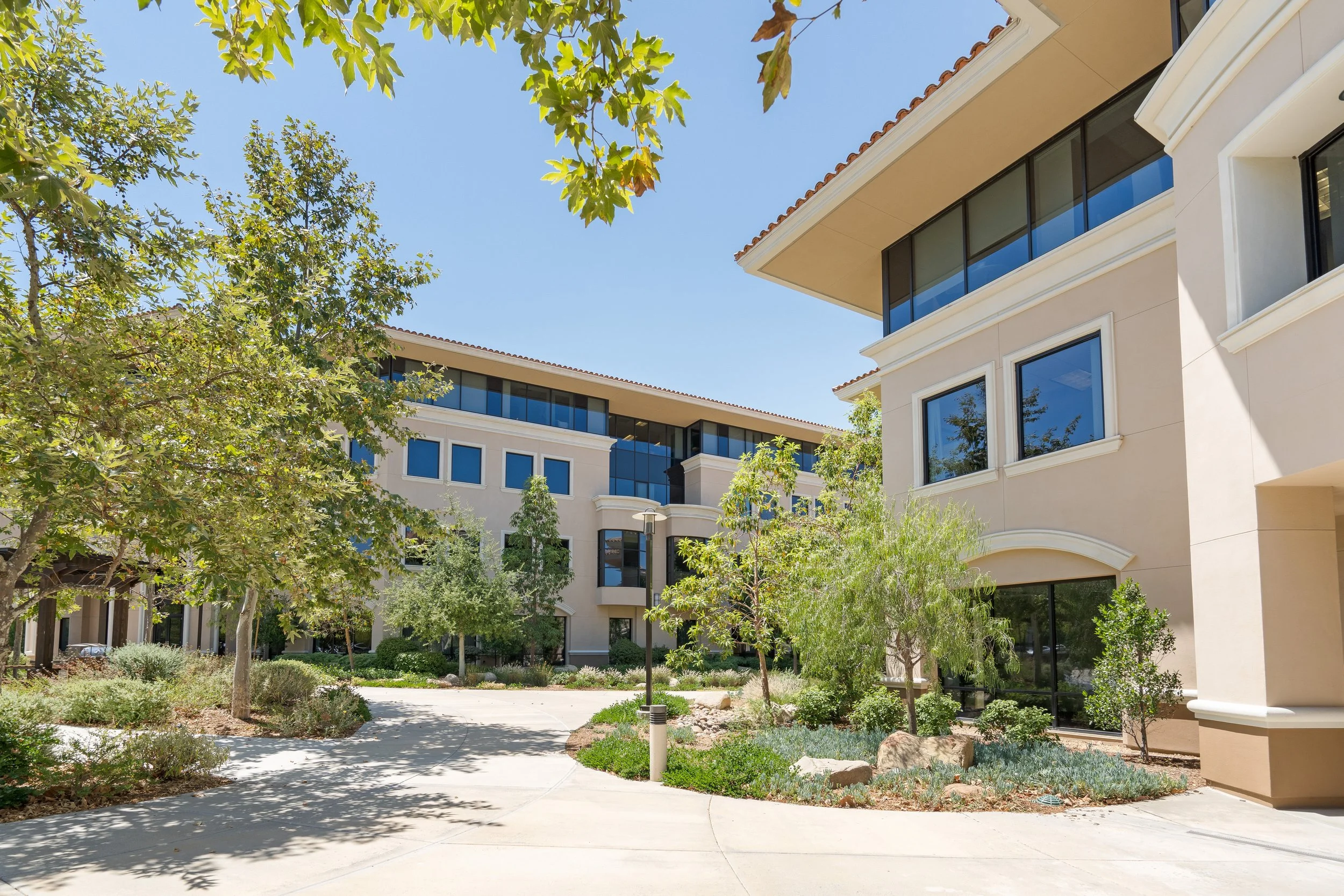 Photo of a modern residential apartment complex with beige walls, large windows, and balconies, set against a bright blue sky, with landscaped trees and bushes in the foreground.