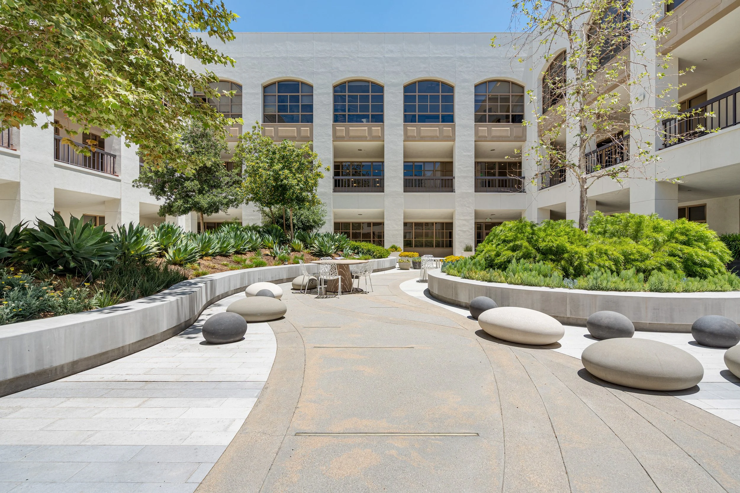 A modern outdoor courtyard with seating, greenery, and multi-story white building in the background.