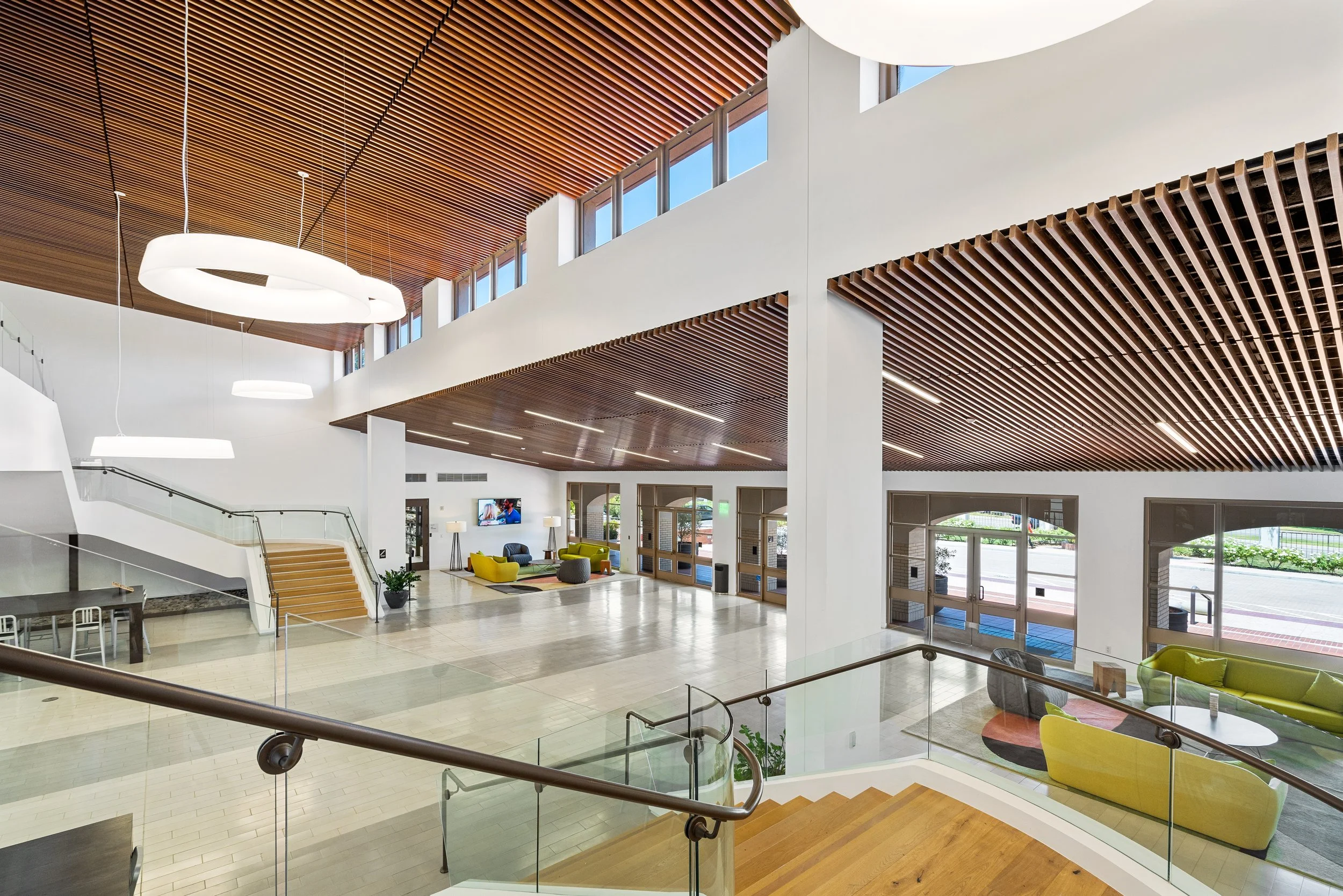Modern hotel lobby with high ceiling, wooden panel accents, and large windows. Features colorful lounge chairs, sofas, and a television.