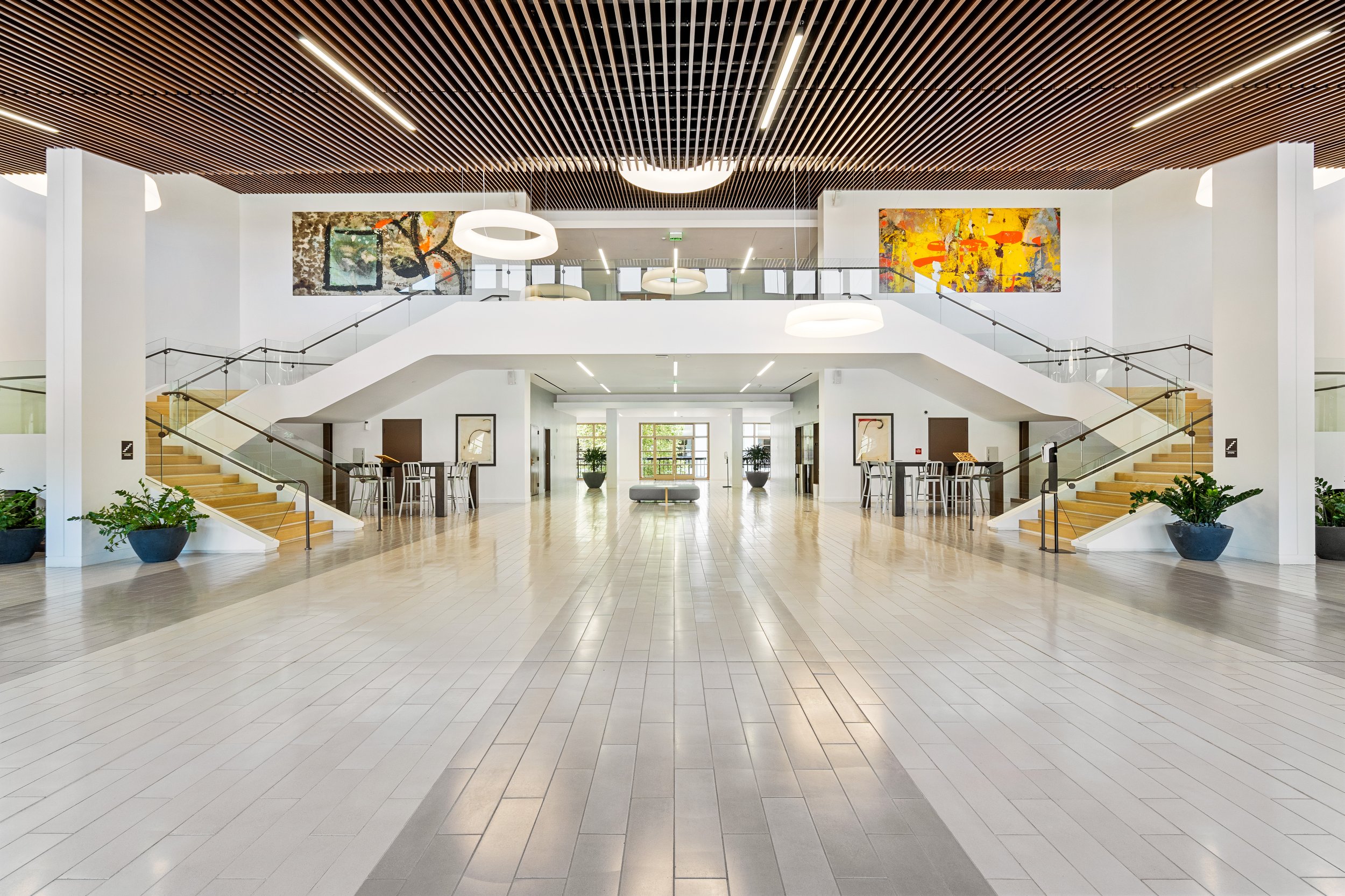 Modern hotel lobby with wooden ceiling, white walls, and a polished tile floor, featuring a staircase on each side leading to an upper level, large abstract paintings, and seating areas with tables and chairs.