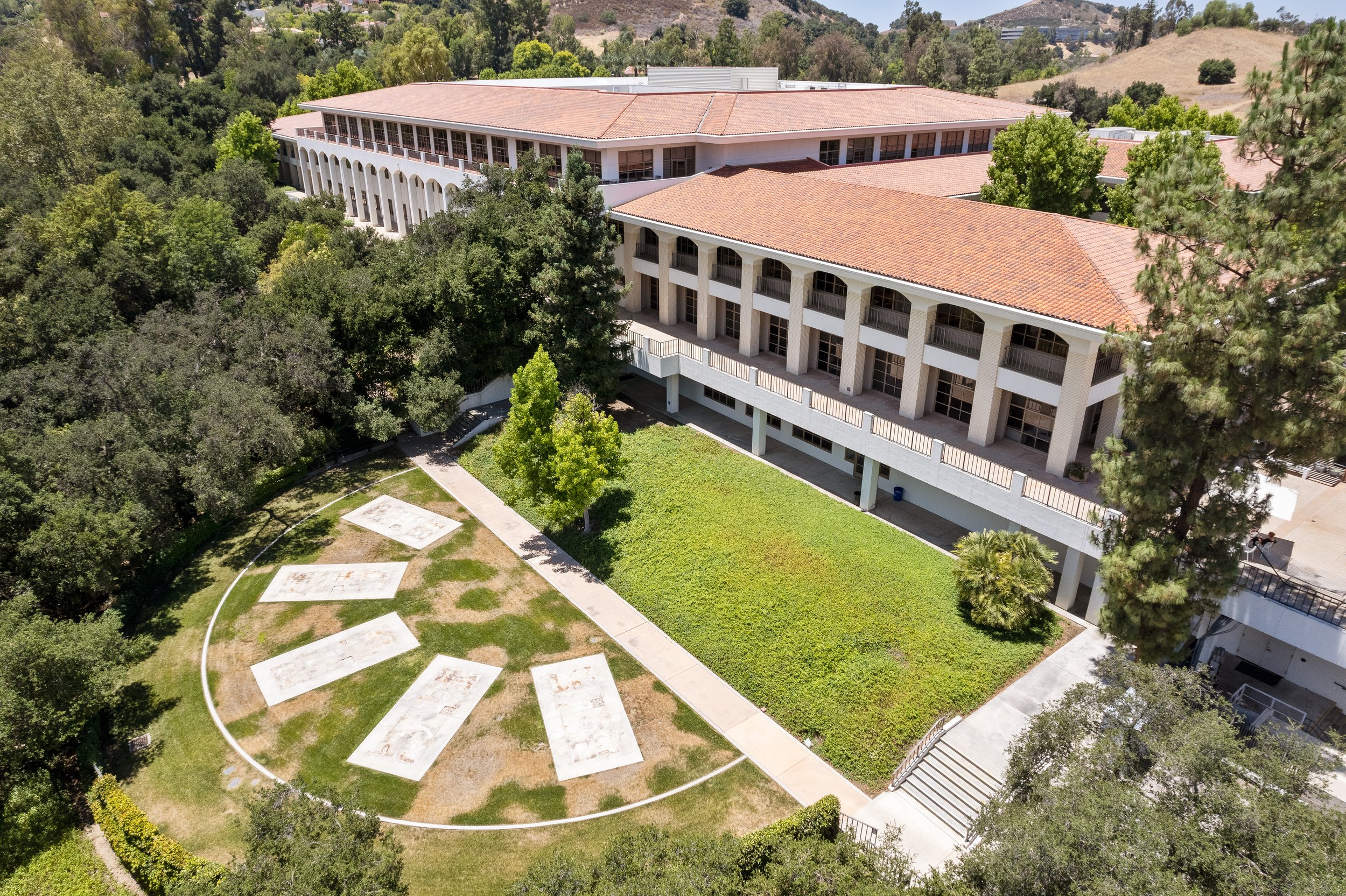  aerial view of a multi-story academic building with red-tile roof, surrounded by trees, with a green lawn and a large outdoor art installation or mural on the ground in front.