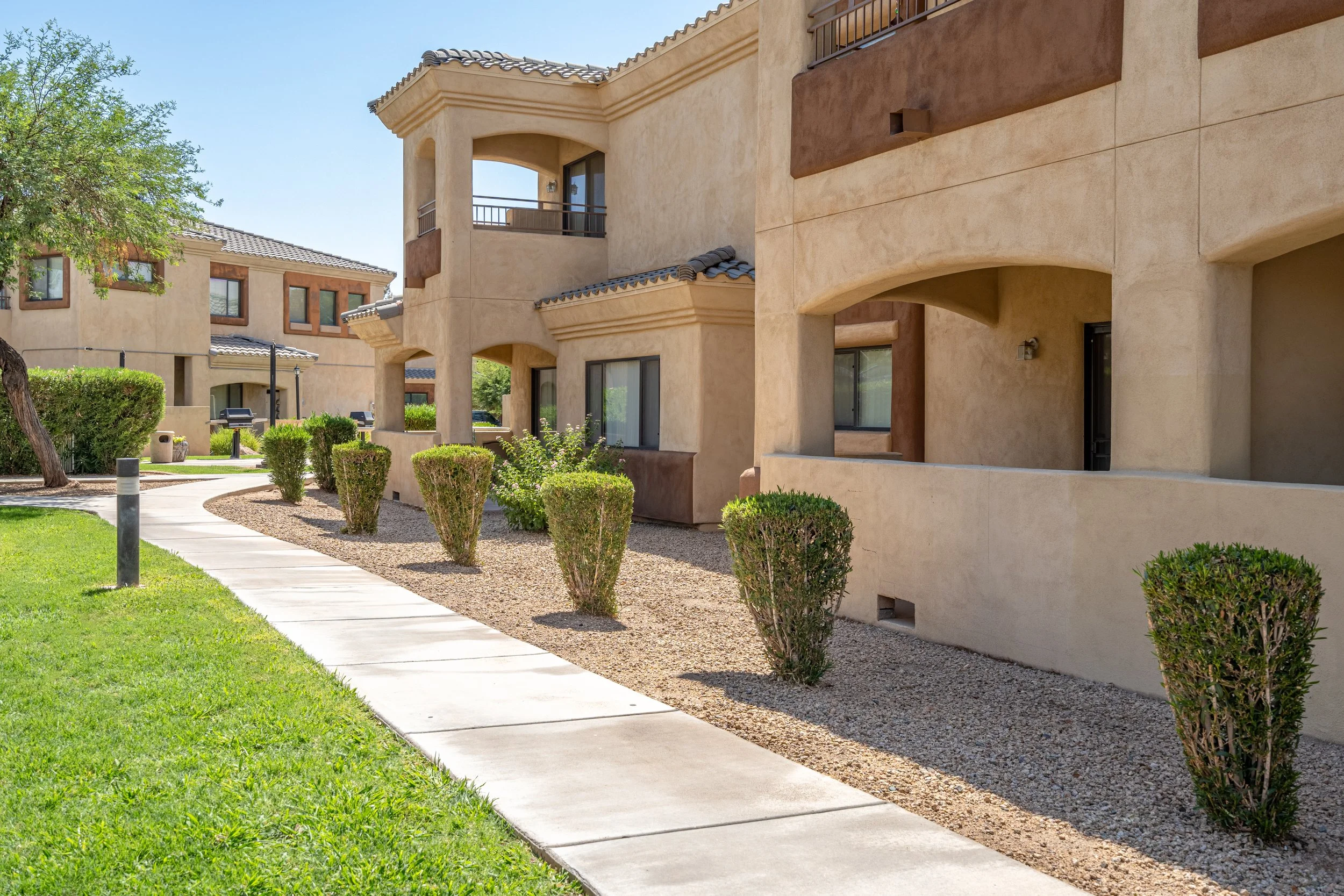 A walkway in front of a beige apartment complex with balconies and green shrubs along the path, sunny sky.