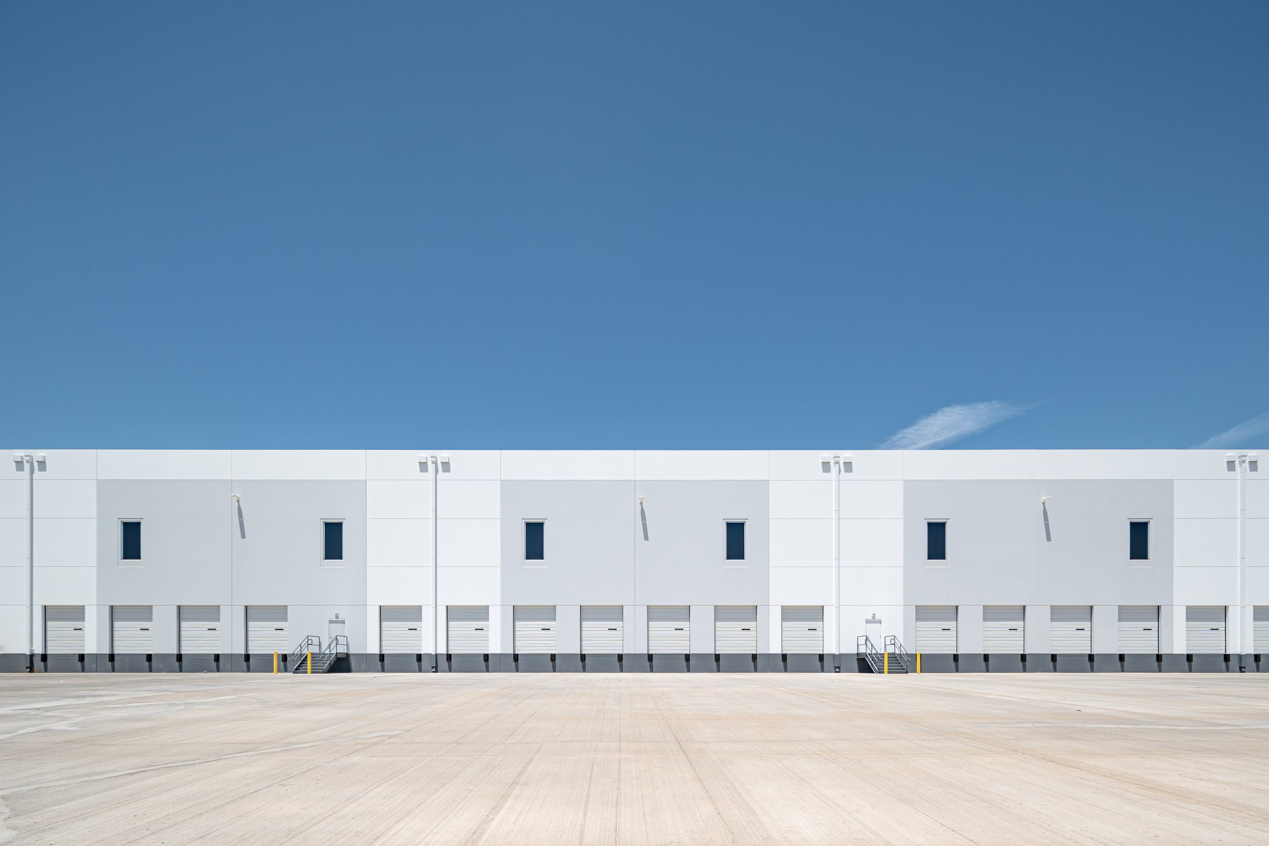 White industrial warehouse building with multiple garage doors and small windows, set against a clear blue sky.