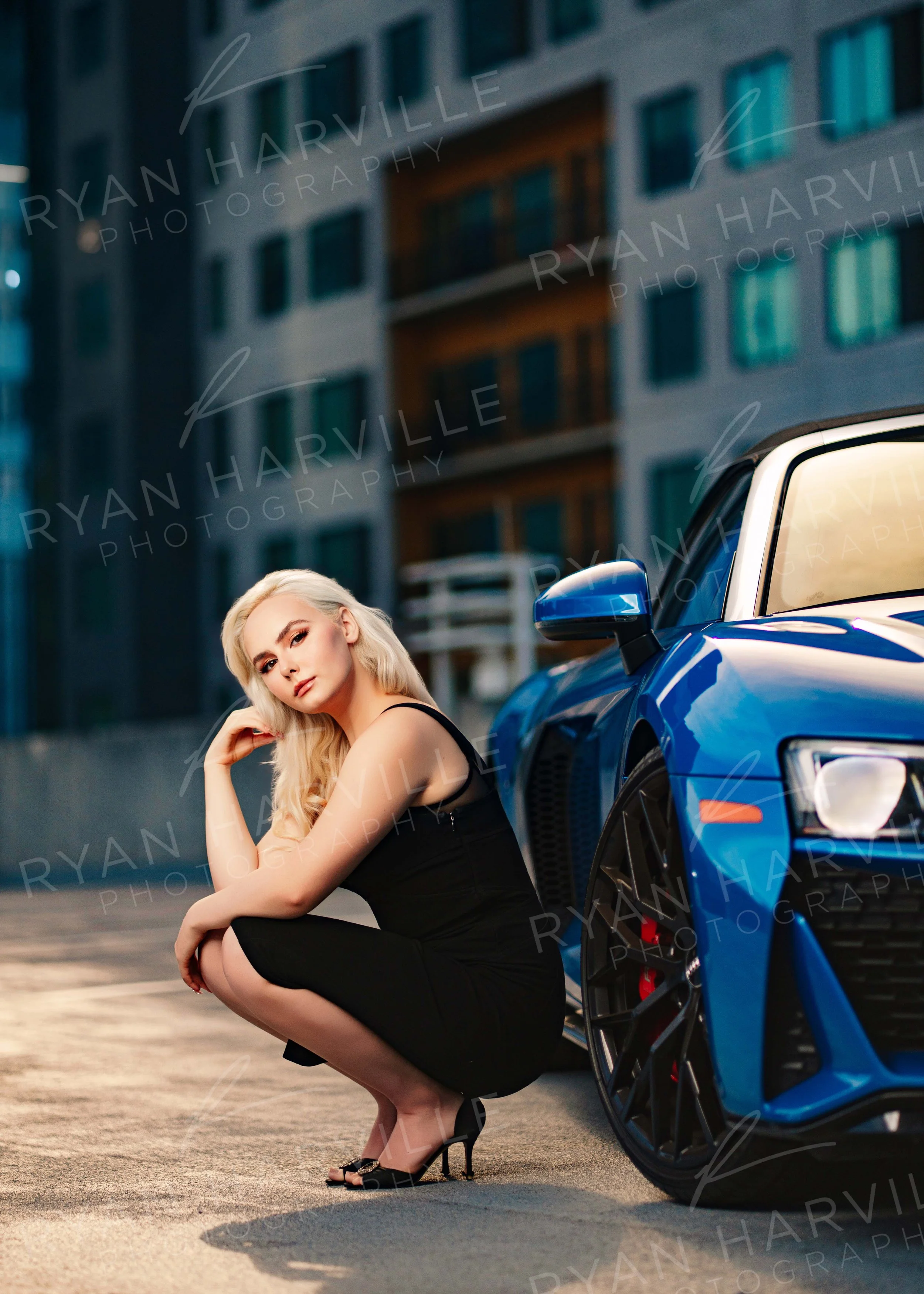 A woman with blonde hair in a black dress crouches next to a blue sports car in an urban setting at dusk.