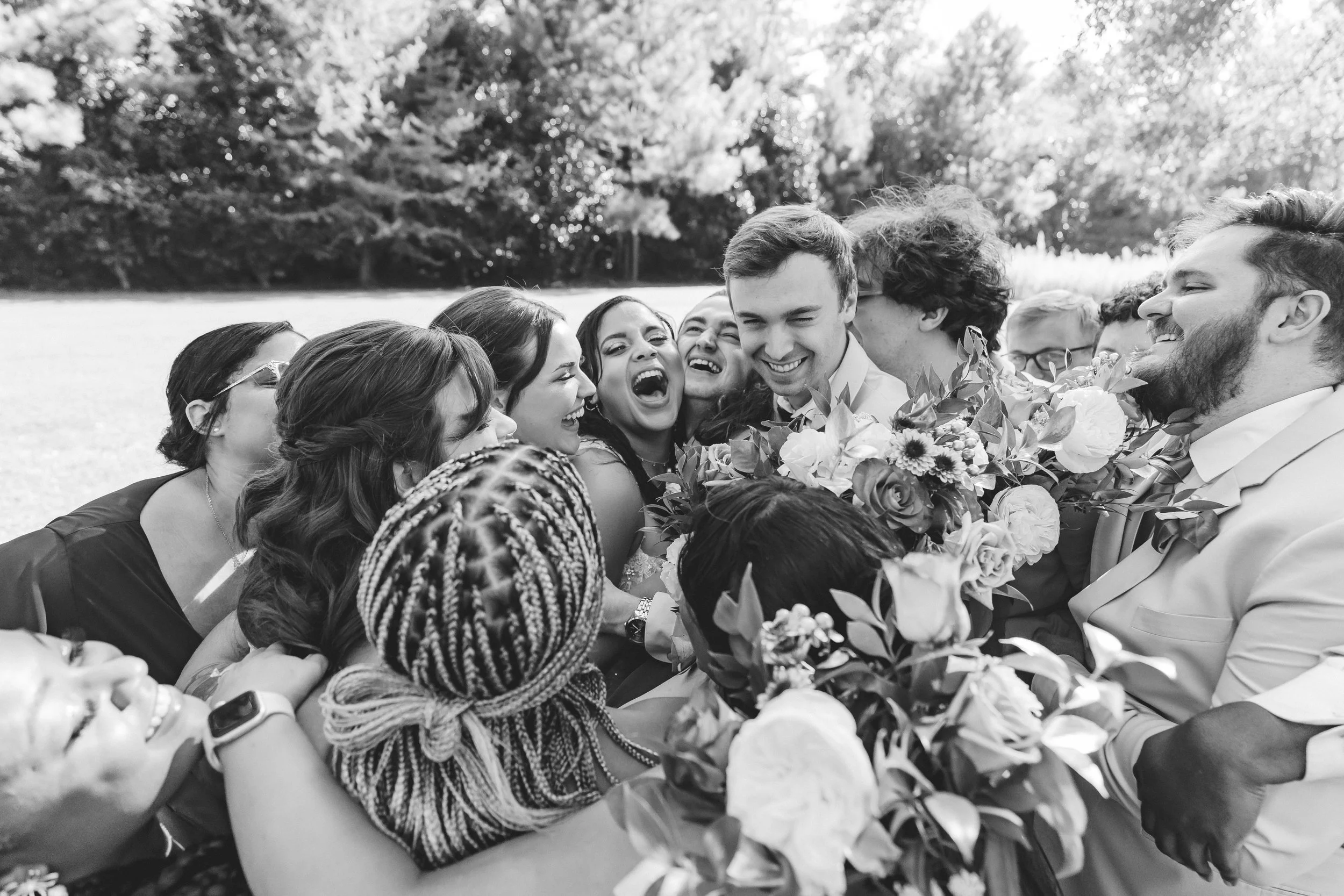 Black and white photo of a group of people smiling and laughing, gathered closely together outdoors, holding bouquets of flowers.