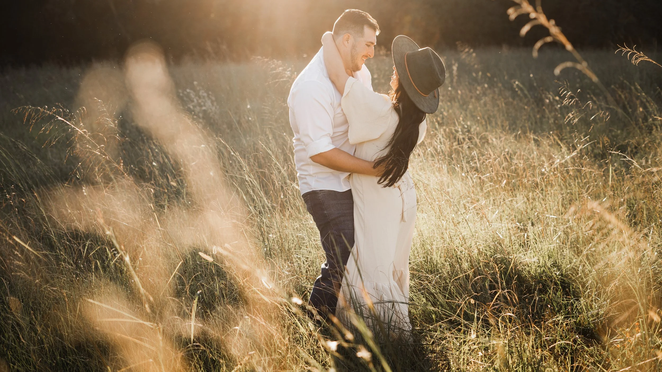 A couple dancing together in a sunlit field of tall grass, with the man holding the woman close and both smiling, during golden hour.