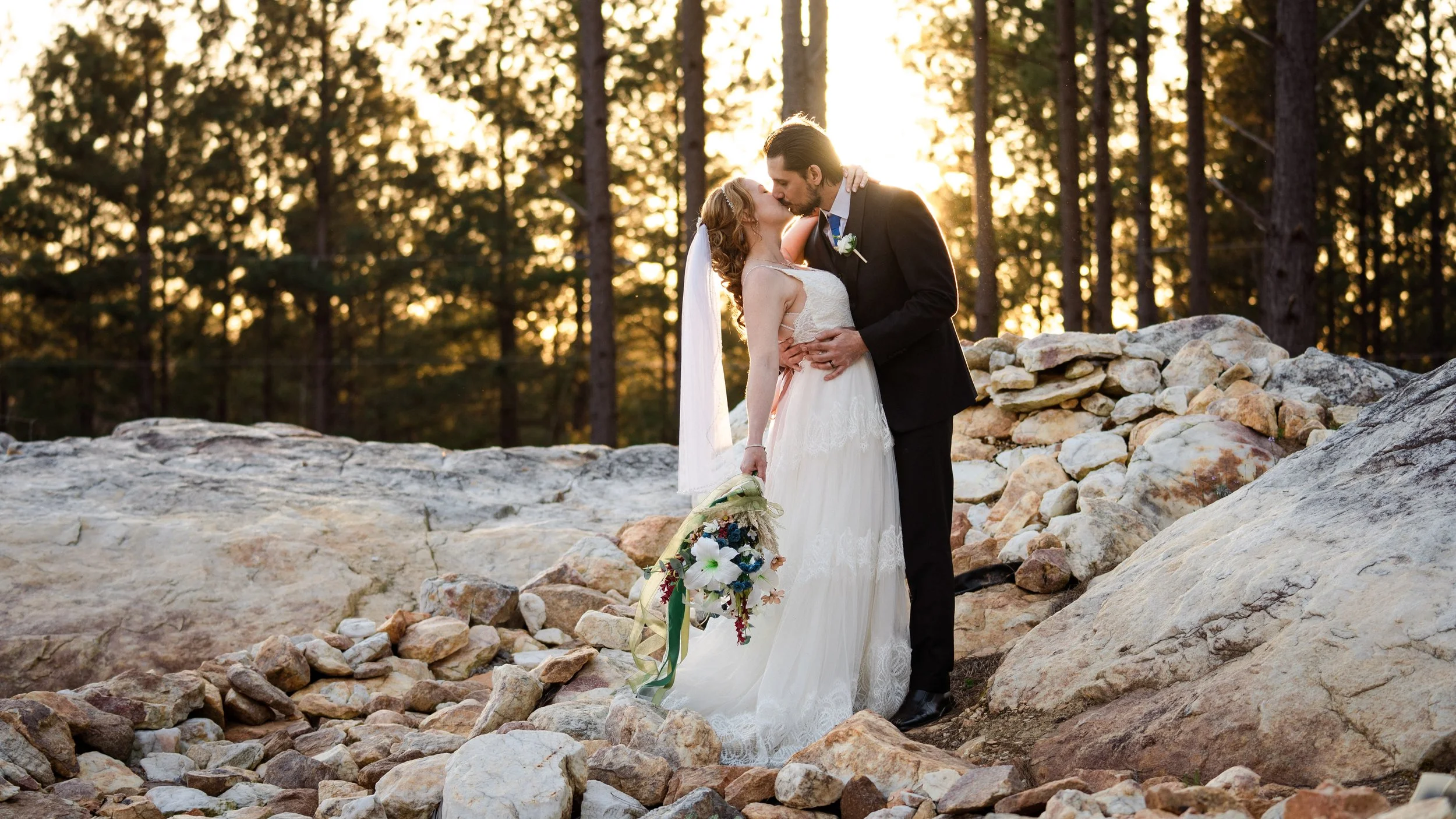 West Georgia bride and groom sharing a kiss during their outdoor wedding at sunset, standing on rocky terrain with trees in the background.