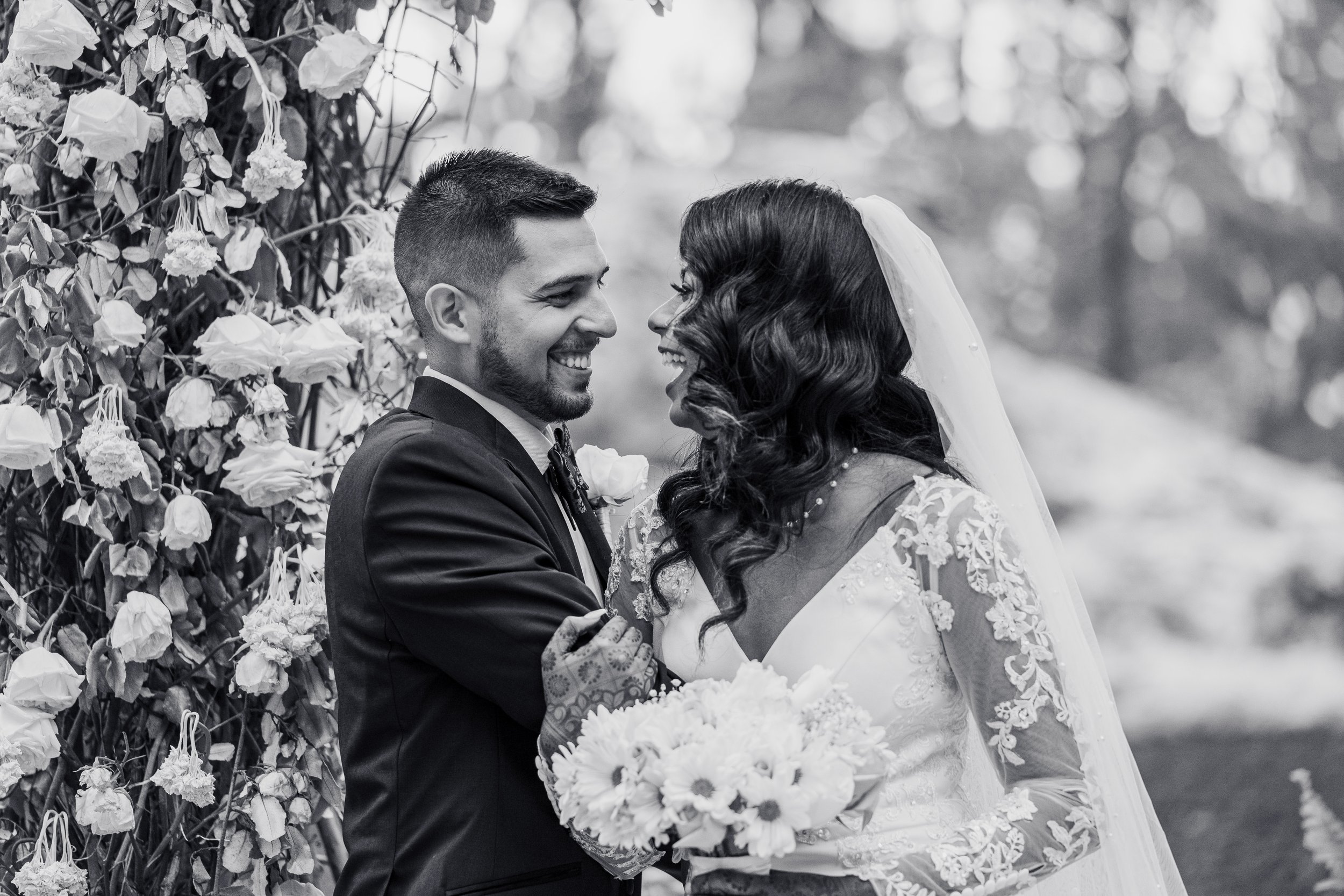 A black and white photograph of a bride and groom smiling at each other, holding hands, with a floral backdrop.