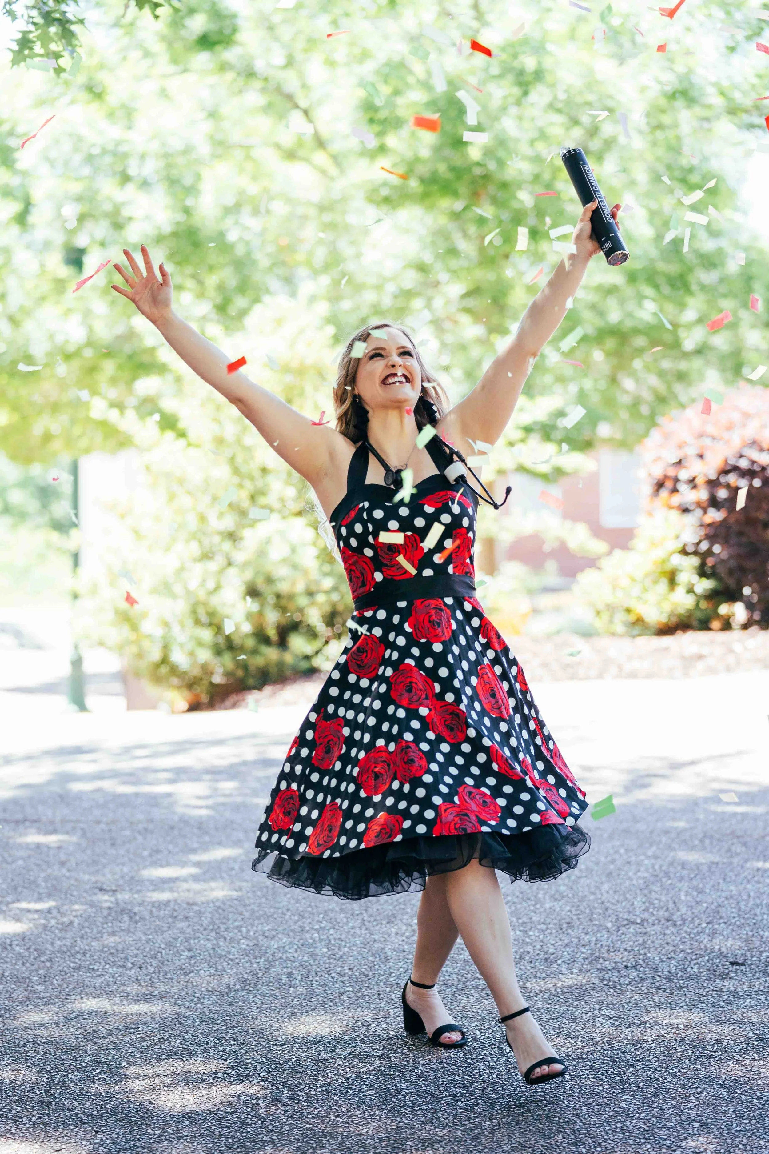 Atlanta college grad in a black dress with red roses and white polka dots celebrating outdoors, throwing confetti, holding a diploma, with arms raised and smiling.